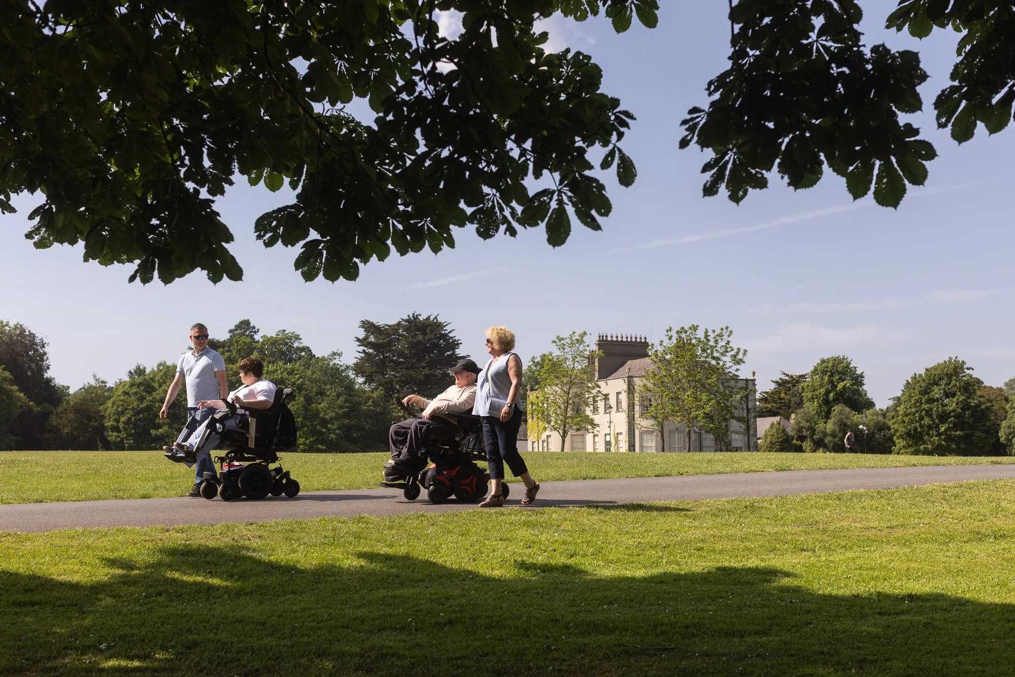 Four elderly people, two in wheelchairs and two standing, are walking along a paved path in a park on a sunny day, with lush green trees and a large building in the background.