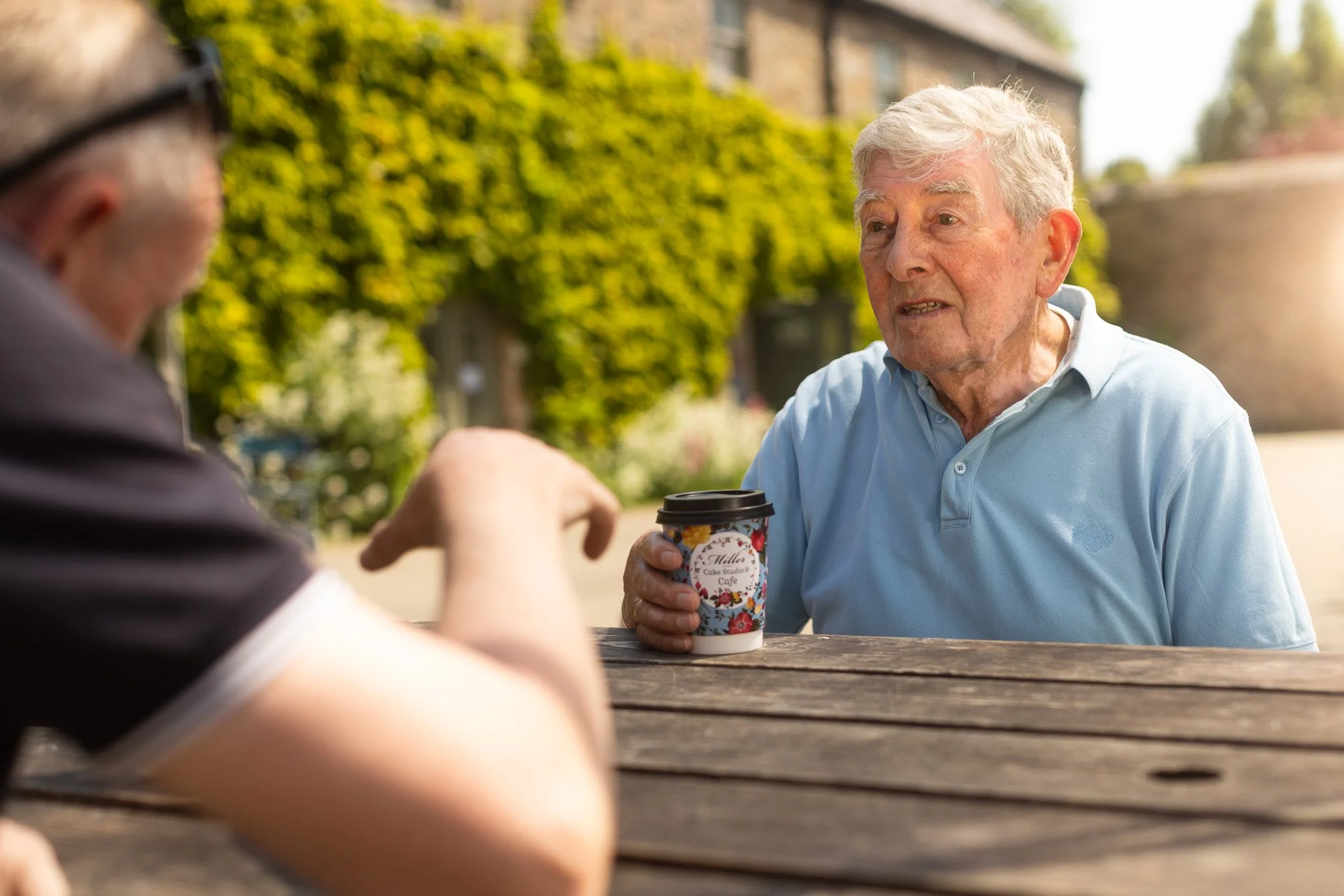 Two elderly men sitting at a wooden picnic table outdoors, one holding a colorful flower-themed coffee cup, engaging in conversation on a sunny day with lush greenery in the background.