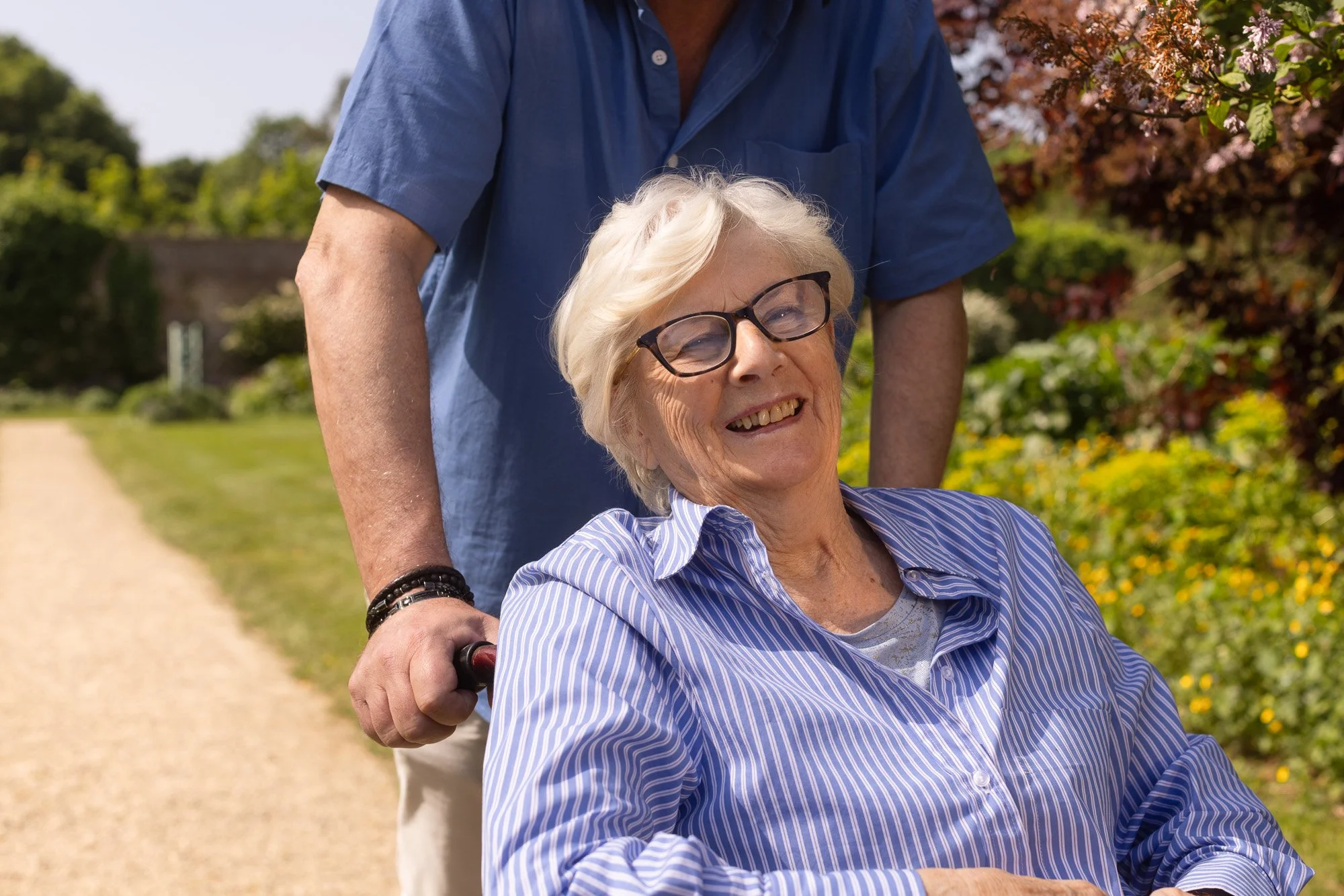 An elderly woman with glasses smiling as she sits in a wheelchair outdoors on a sunny day, with a man pushing her from behind along a dirt path surrounded by blooming trees and greenery.