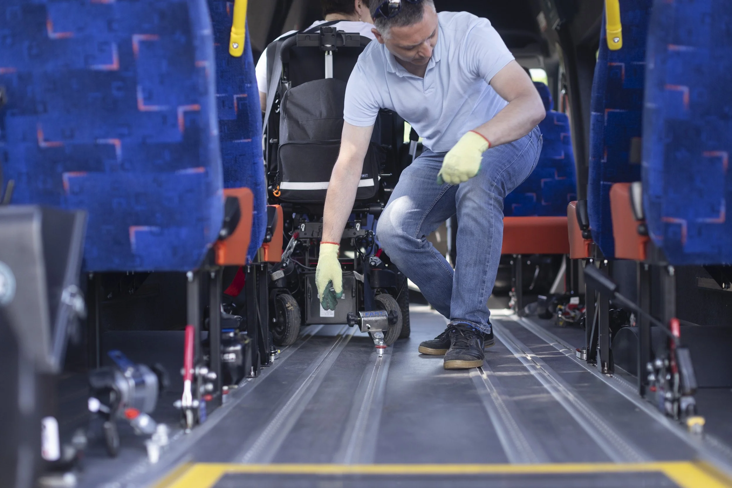 A man in a white shirt and jeans kneels next to a wheelchair inside a bus, working on the wheelchair's mechanisms. He is wearing yellow gloves and appears to be performing maintenance or repairs in a professional environment.