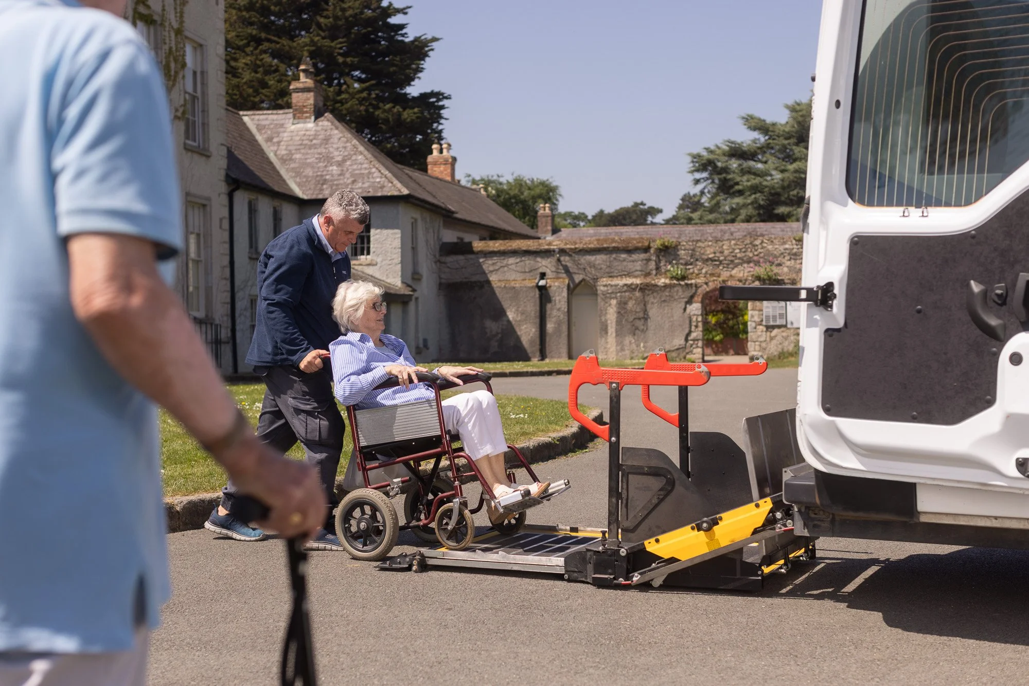 A person being transferred from a wheelchair into a van with a wheelchair lift by medical staff, with another person partially visible in the foreground.