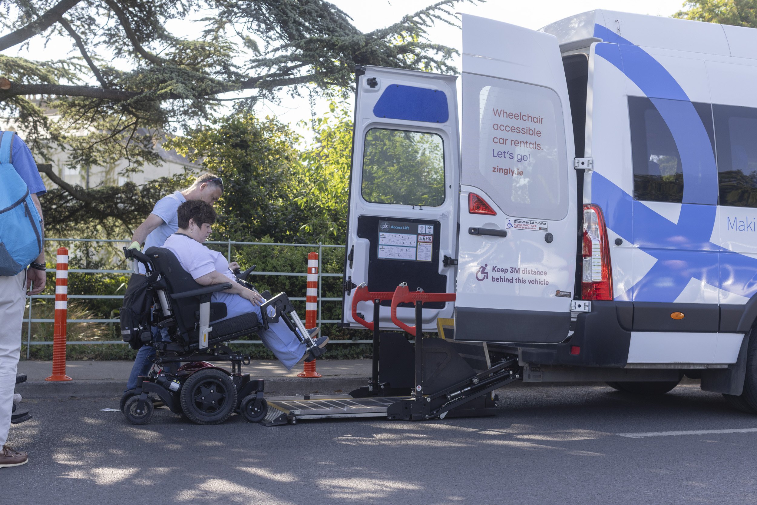A person in a wheelchair is being assisted onto a van with a wheelchair lift. The van has signage indicating it offers accessible car rentals. Two other individuals are nearby, and there are orange safety cones and plastic barriers around the area.