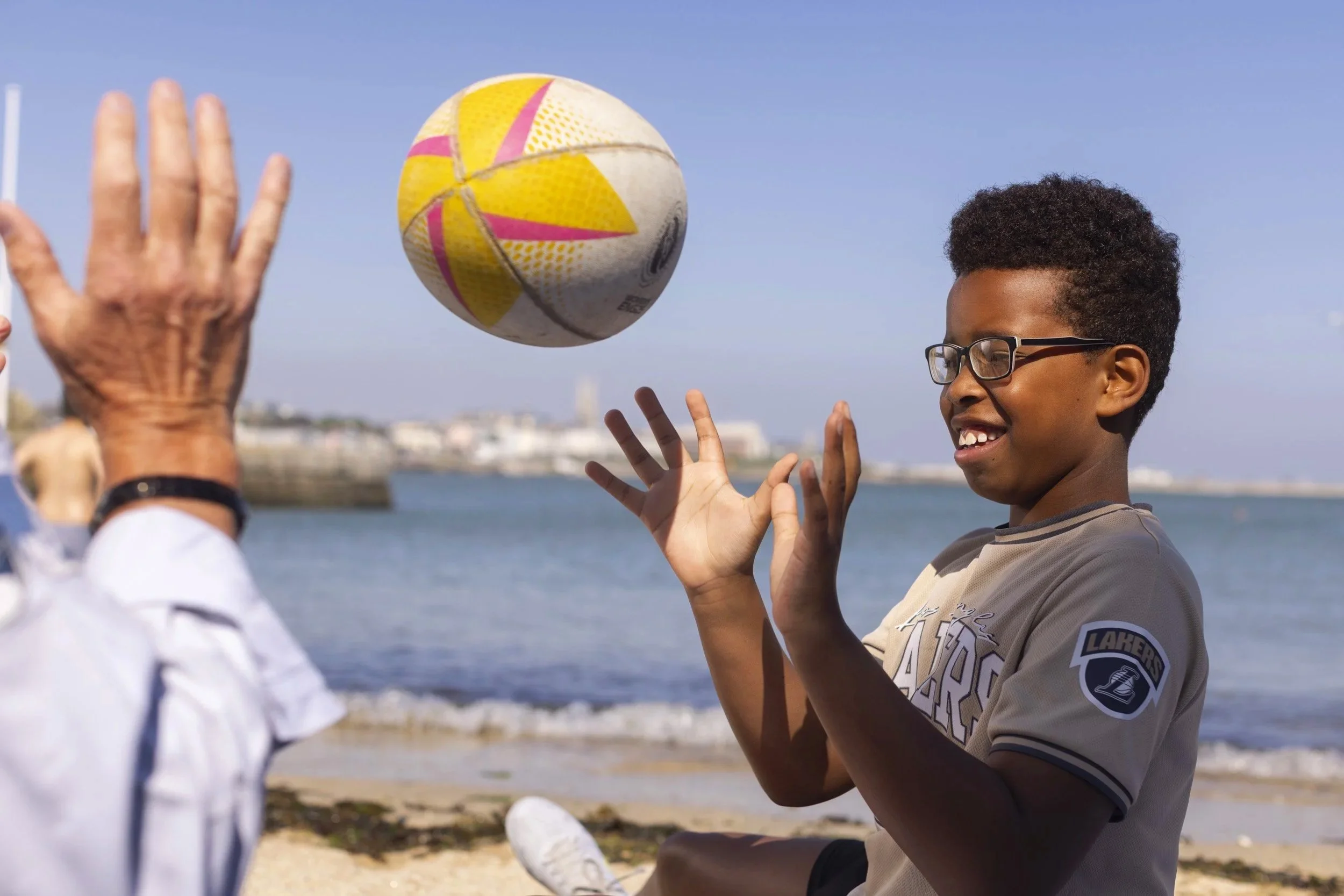 A young boy playing beach volleyball outdoors with a smiling person whose face is not visible, volleyball in mid-air, ocean or large body of water in the background, clear blue sky.