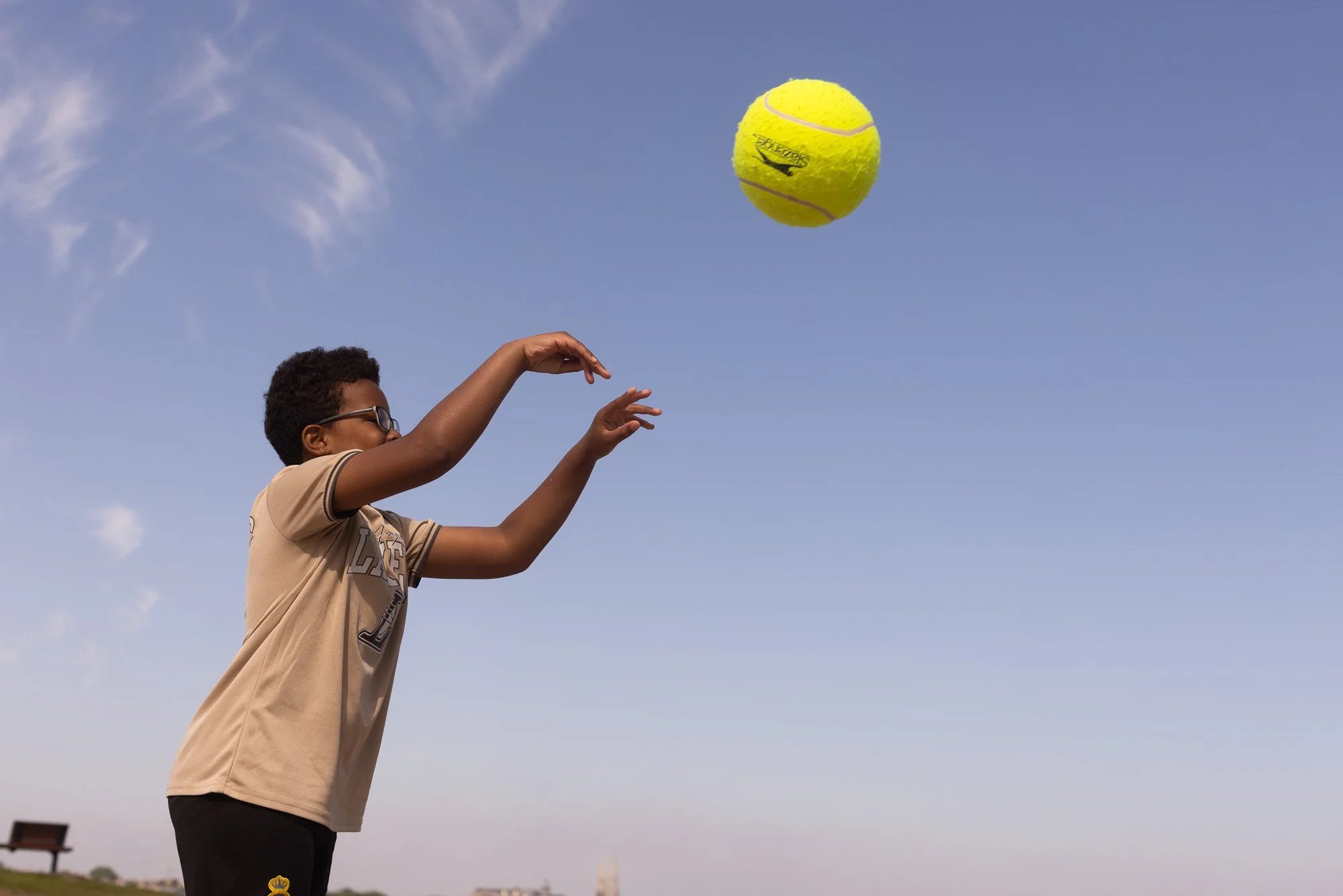 A young person with short curly hair and sunglasses tossing a bright yellow tennis ball into the air outdoors against a clear blue sky.