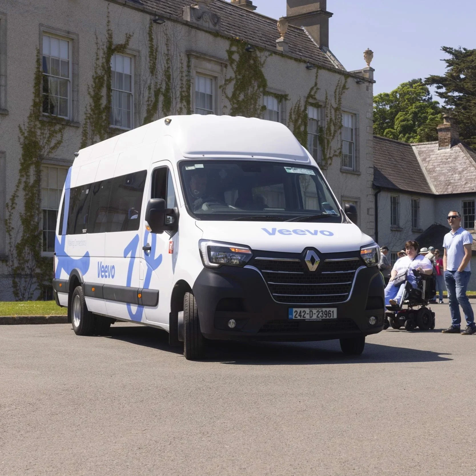 A white van with the word "Veevo" written on it in blue, parked on a street in front of a large cream-colored building with ivy growing on the walls. Several people are walking nearby, including a man in sunglasses, a woman pushing a stroller, and a child in front of the building.
