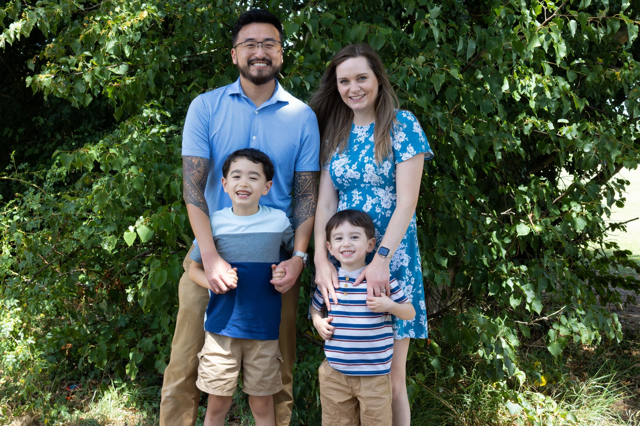 A diverse family of five smiling outdoors in front of a leafy green bush, with two boys, a woman, a man, and a young girl.