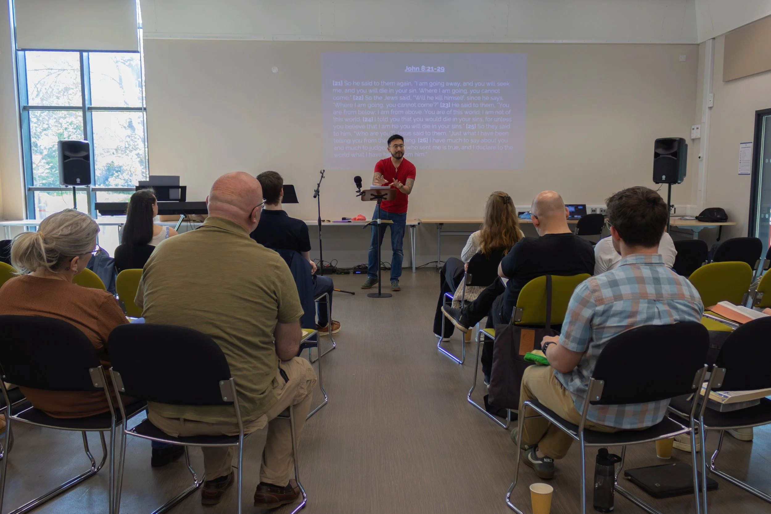 A man in a red shirt giving a presentation to an audience in a bright room with large windows. The presentation slide displayed on the wall shows a scripture passage from John 8:21-29.