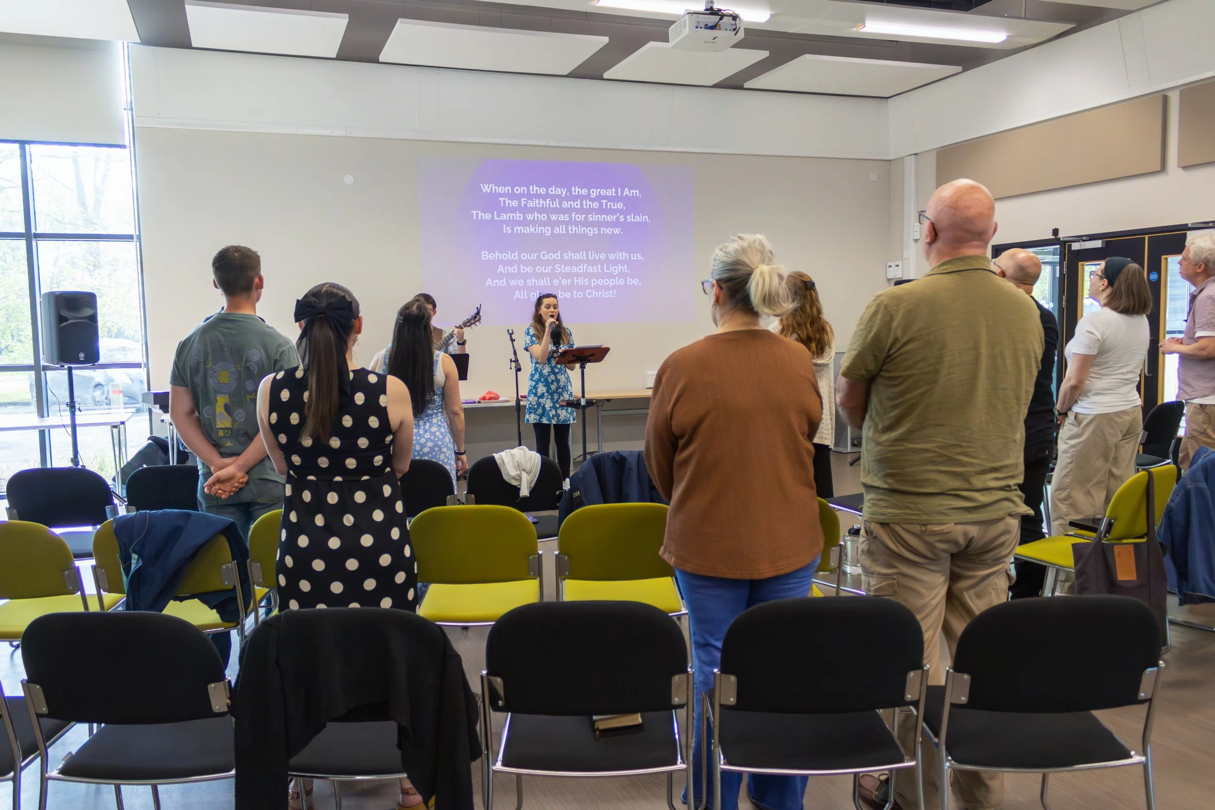 People standing and facing a presentation screen in a modern conference room with large windows, some holding guitars, listening to a woman singing with lyrics projected on the screen behind her.