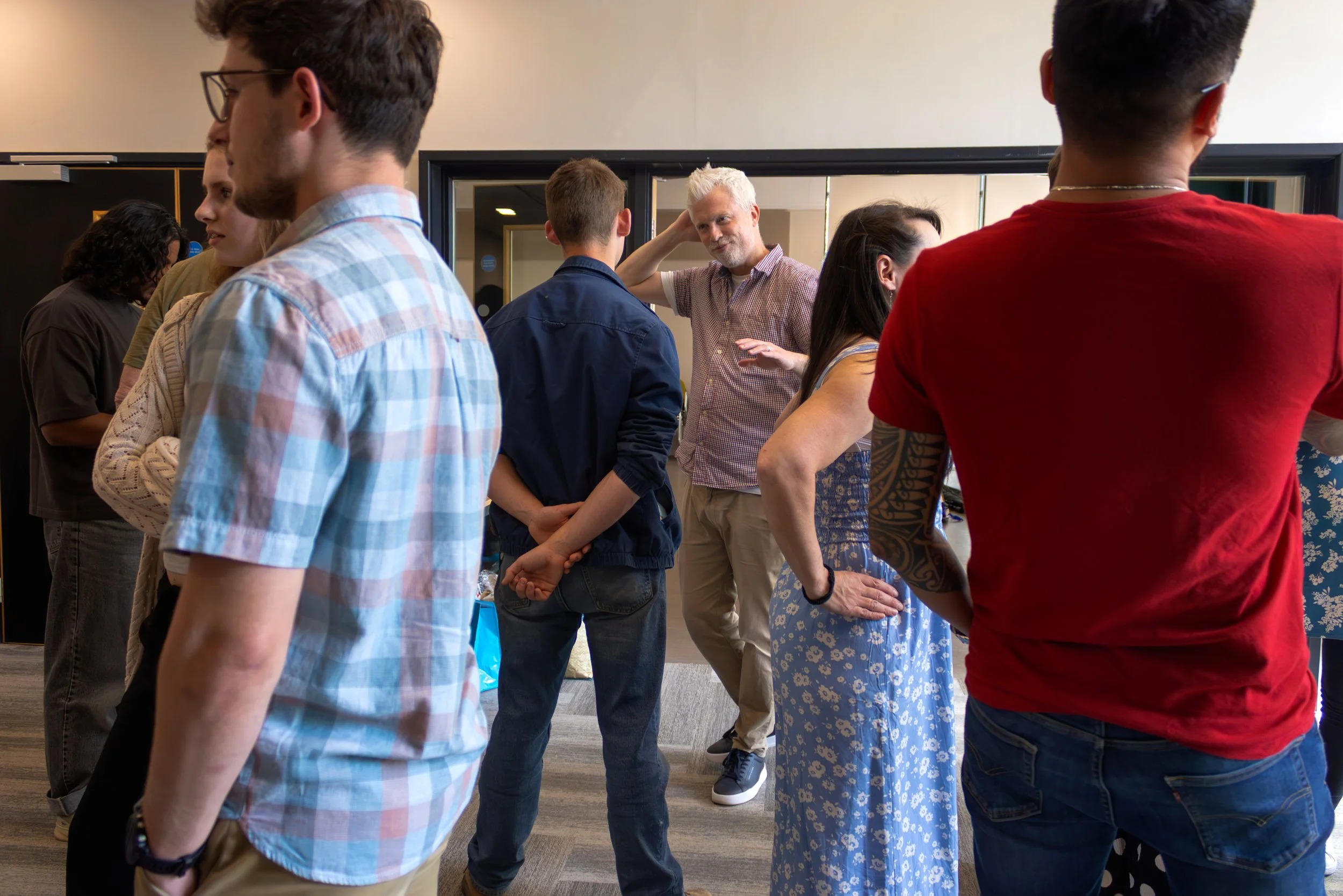 A group of diverse people standing in a line in an indoor setting, engaging with a person with white hair who appears to be leading or instructing them.