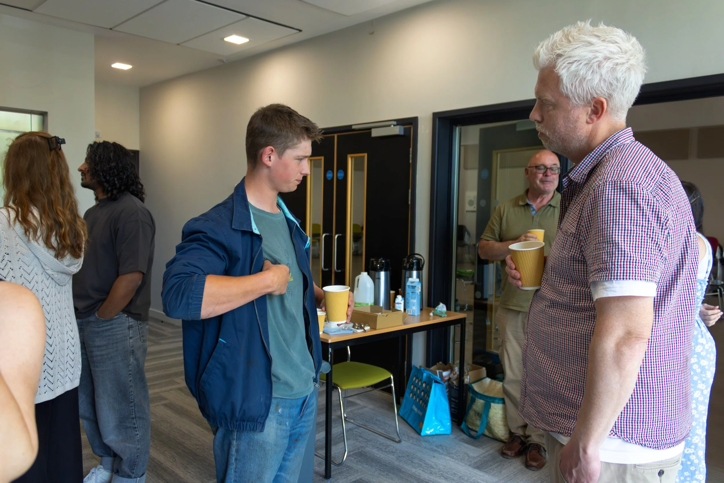 People engaged in conversation at an indoor social gathering, with some holding paper cups and a table with beverages in the background.