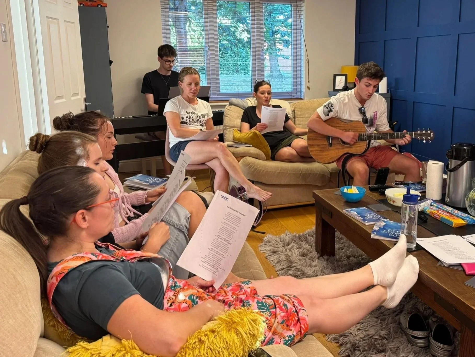 A group of people in a living room, some sitting on a beige couch reading printed scripts, others performing a song with one person playing guitar. One person is lying with legs raised, reading a script, while a young man is playing guitar. The room has a coffee table with snacks, a large window with blinds, and a blue accent wall.