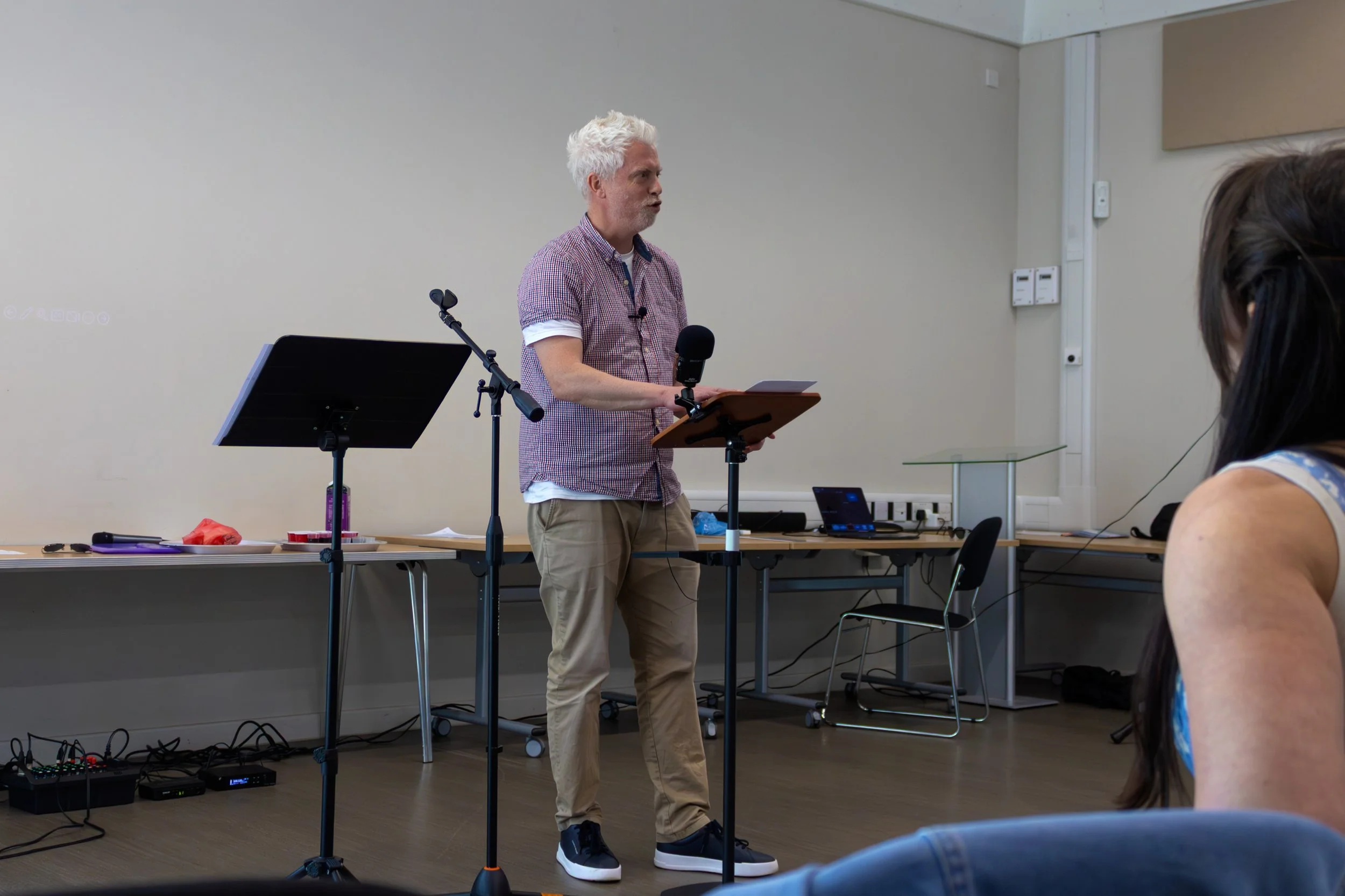 A man with white hair and a beard, wearing a checkered shirt and beige pants, is standing and speaking into a microphone in a classroom or conference room. There is a music stand and various electronic devices on a table beside him. A woman with dark hair and a sleeveless top is sitting and listening to him.