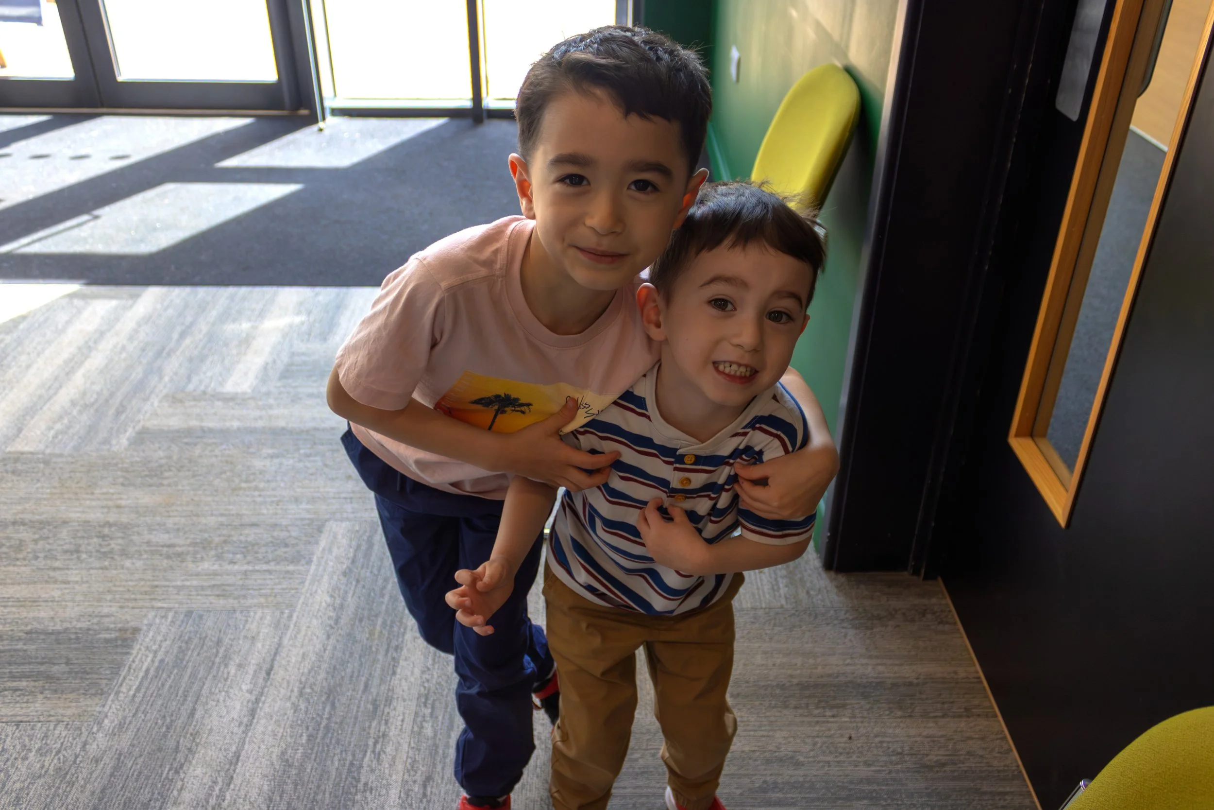 Two young boys hugging and smiling at the camera indoors near glass doors, with sunlight casting shadows on the floor.