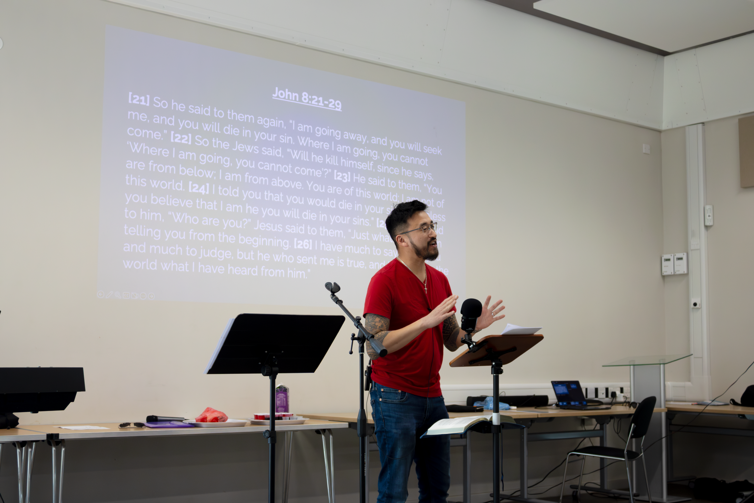 A man with tattoos on his right arm and glasses, wearing a red shirt, standing and speaking in front of a large screen displaying a Bible passage, with a microphone and music stand nearby in a classroom or conference room.