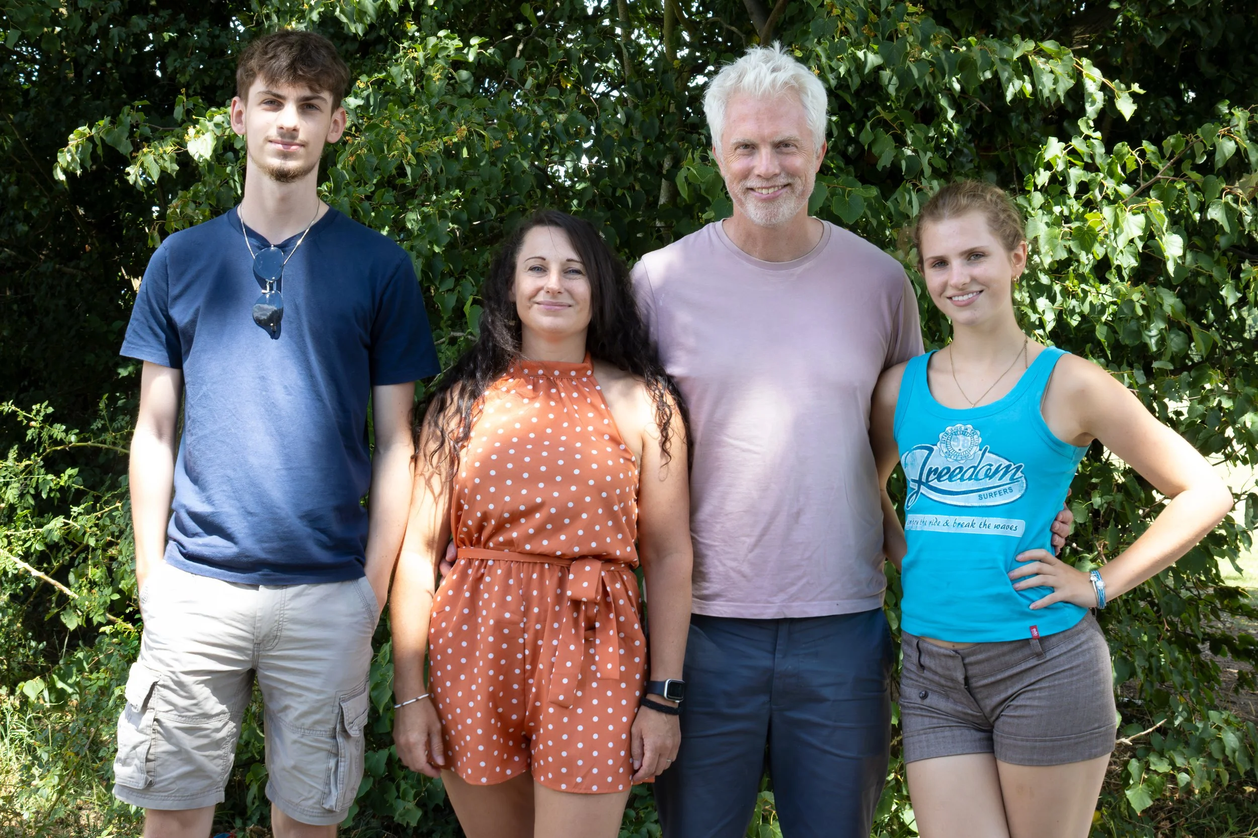 A group of four people standing outdoors in front of green leafy trees, smiling at the camera. They include a young man in a blue shirt and sunglasses hanging from his shirt, a woman in a orange polka dot dress, a man with white hair and a purple shirt, and a young woman in a blue tank top.
