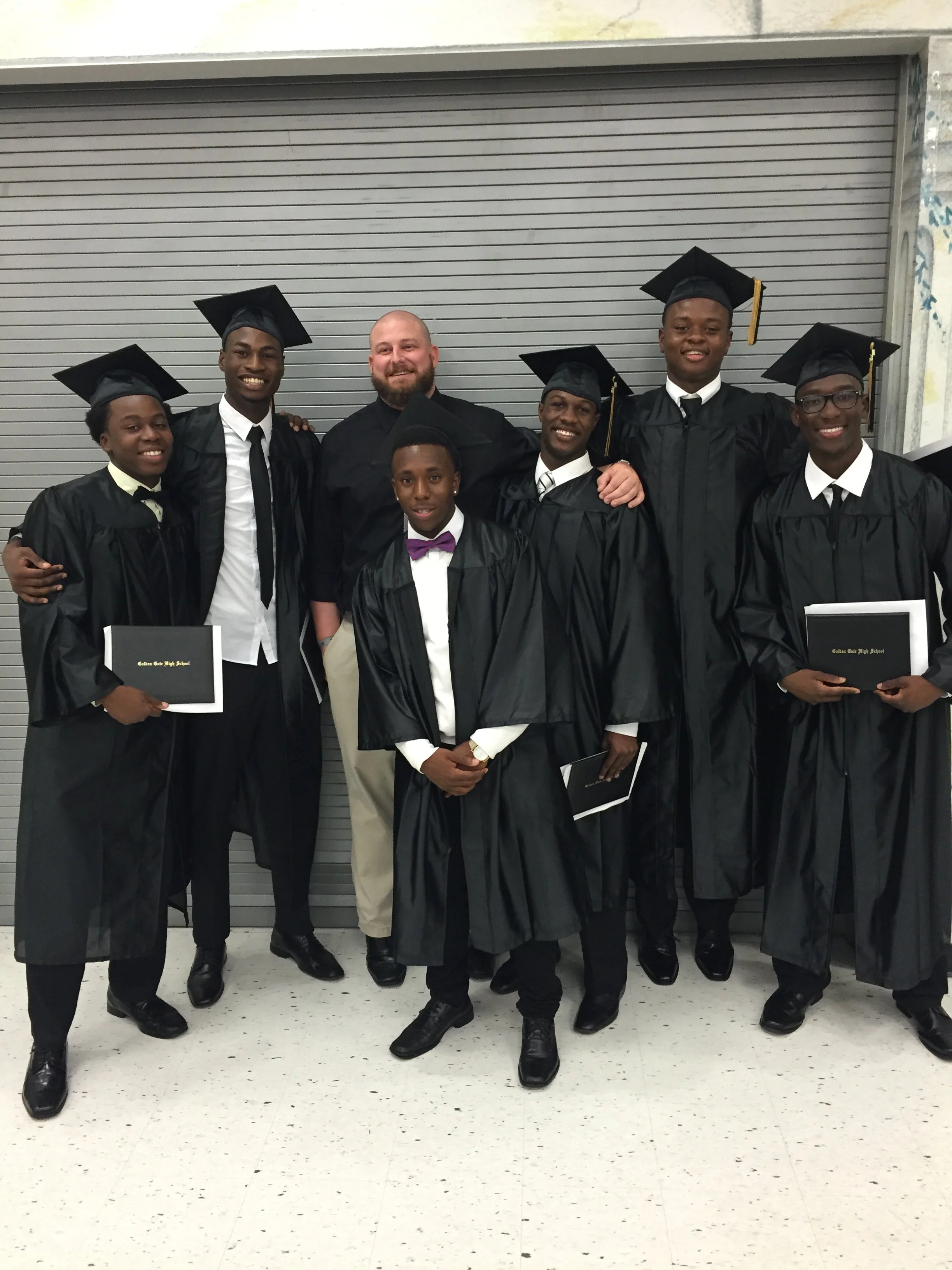 Group of seven young men and one adult man wearing graduation caps and gowns, standing in front of a gray industrial garage door, celebrating their graduation inside a school.