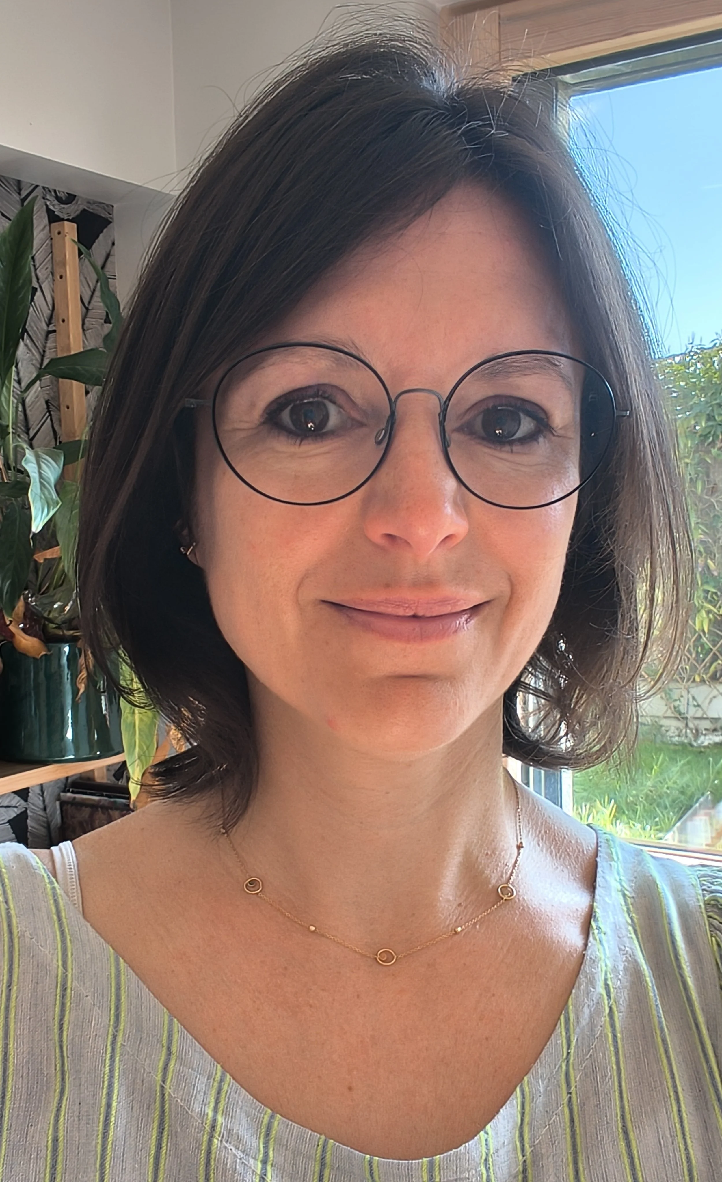 Une femme souriante portant des lunettes rondes, un collier délicat et un haut à rayures vertes et blanches, posant près d'une fenêtre avec lumière naturelle, dans un intérieur décoré de plantes.