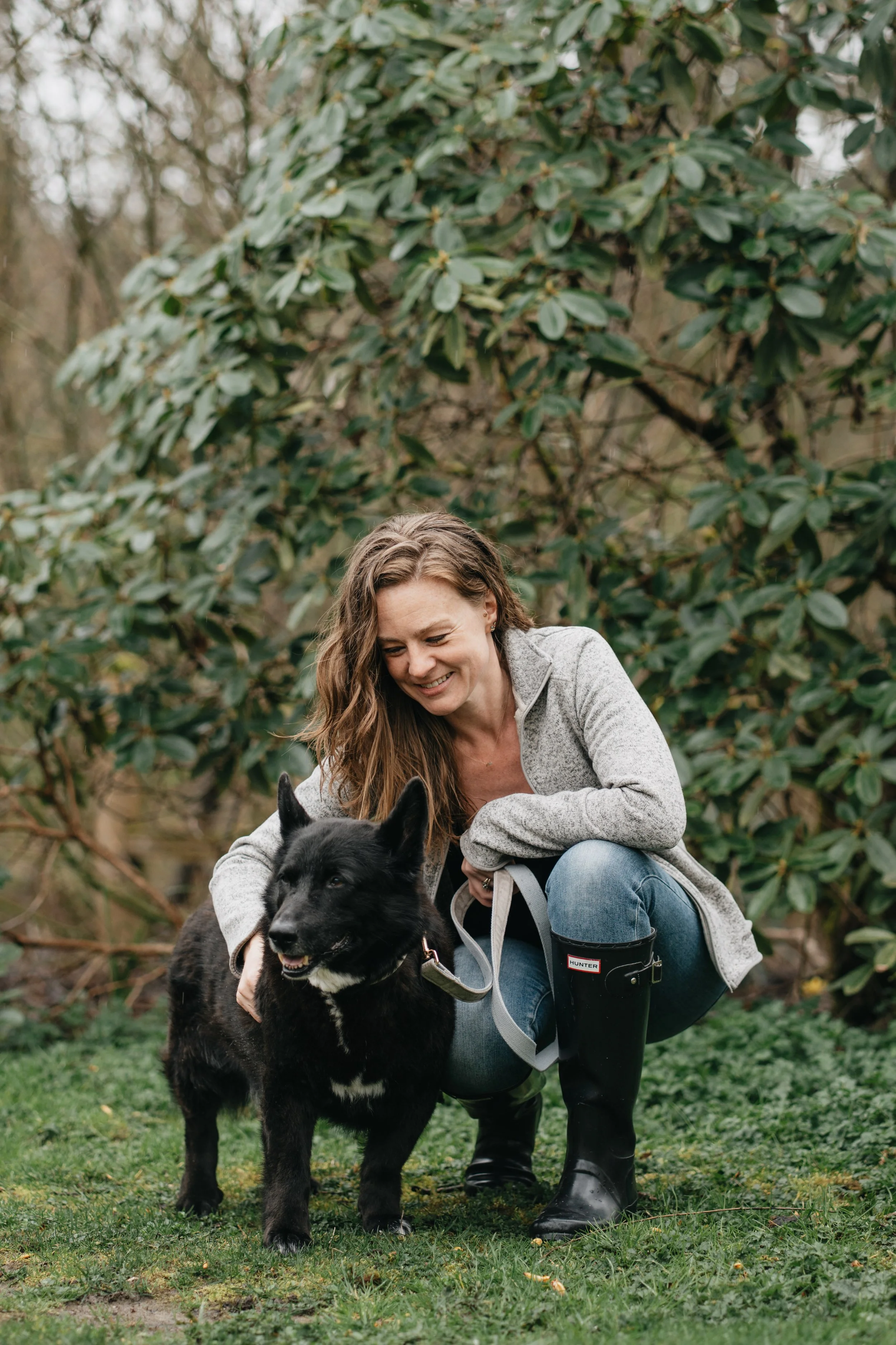 A woman with long brown hair, wearing a grey jacket and rain boots, is crouching on green grass outdoors, petting a black and white dog with pointy ears and a happy expression. The background features green leafy bushes and trees.