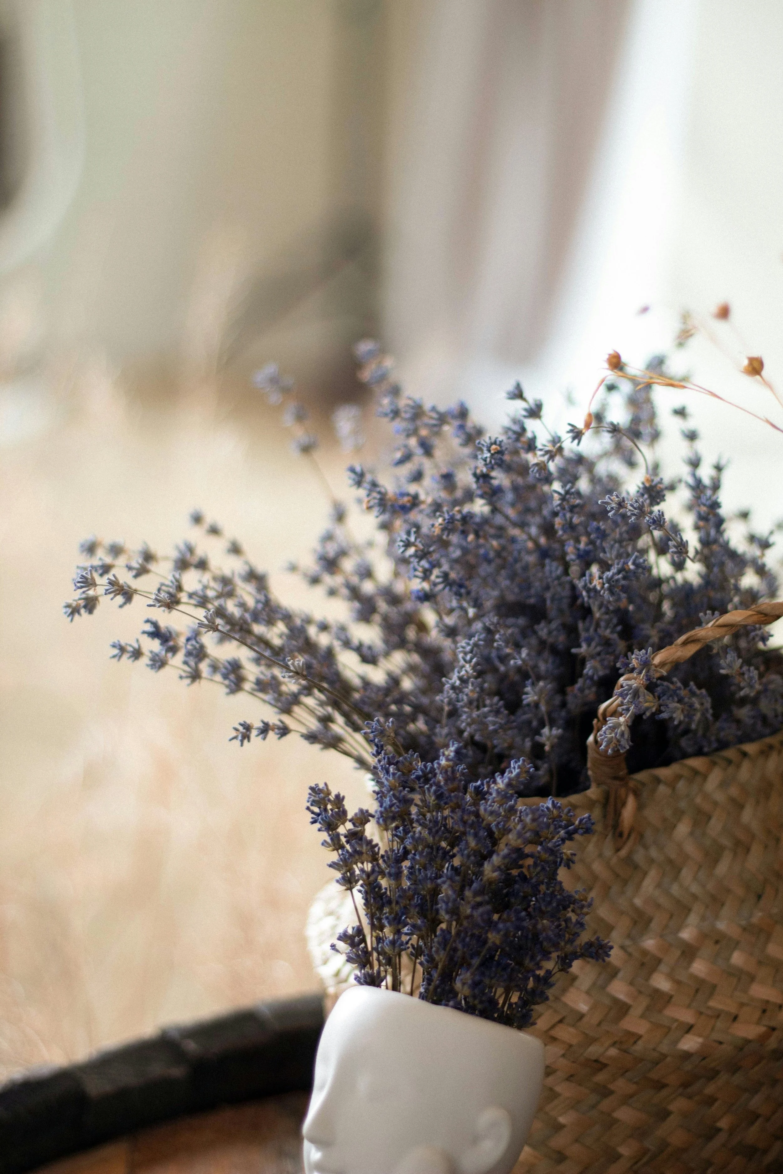 A woven basket with dried lavender flowers on a table, partially obscured by a white ceramic bust of a human head, with a hat on the table.