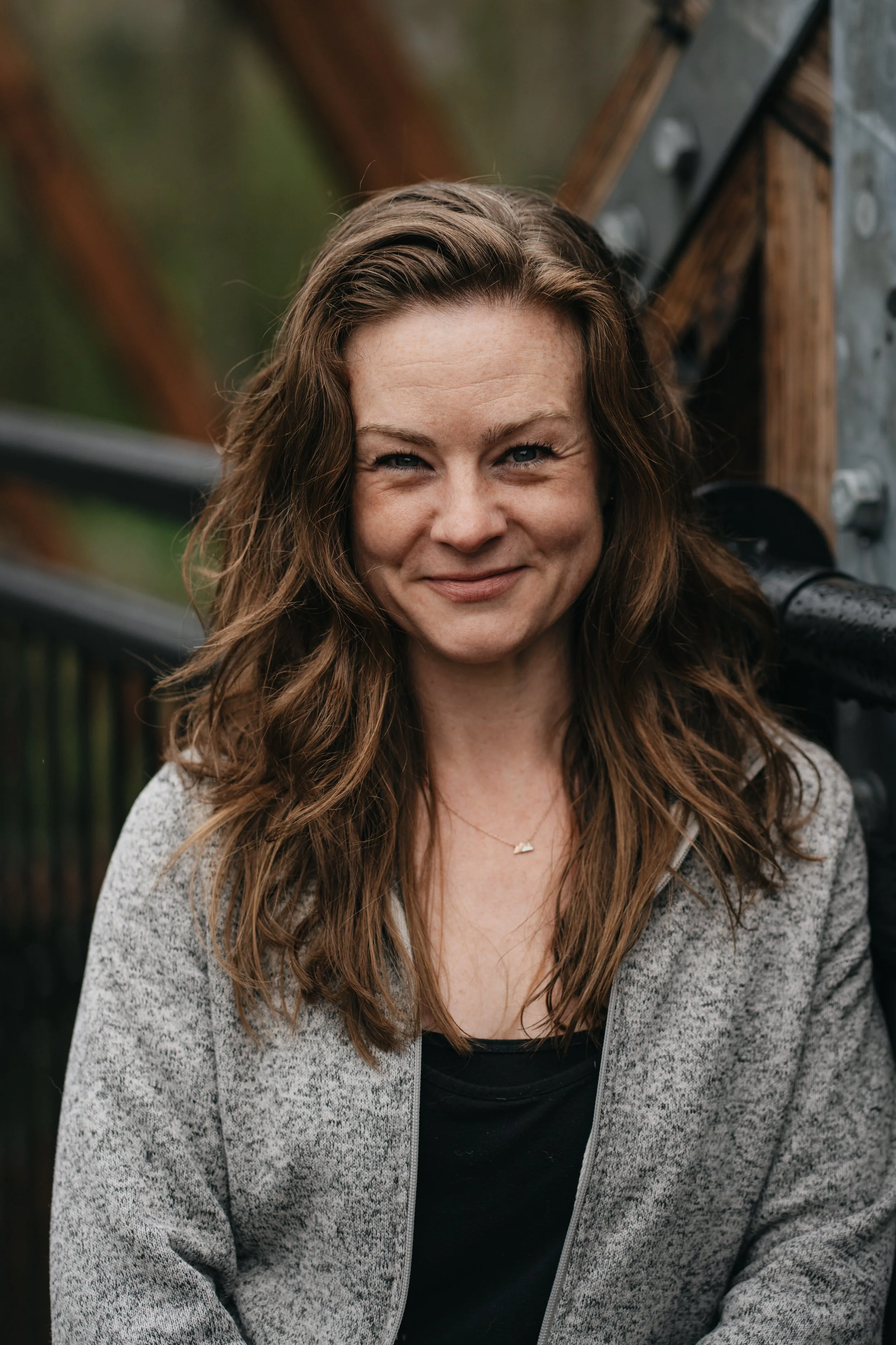Photo of therapist Jenna Schmidt, a woman with long redish brown wavy hair, smiling slightly, wearing a gray zip-up sweater over a black top, standing outdoors with a blurred background of a bridge and greenery.