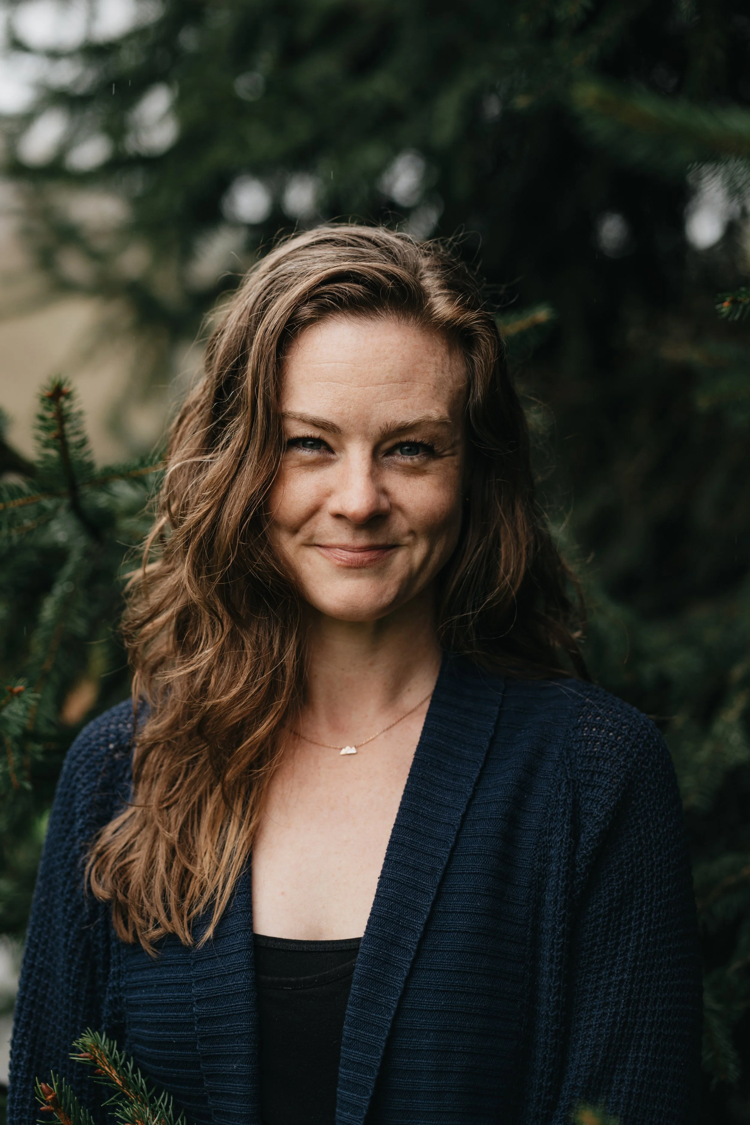 A woman, Jenna Schmidt, with wavy brown hair and blue eyes smiling outdoors, wearing a dark blue cardigan over a black top, with a small necklace, standing among evergreen branches.
