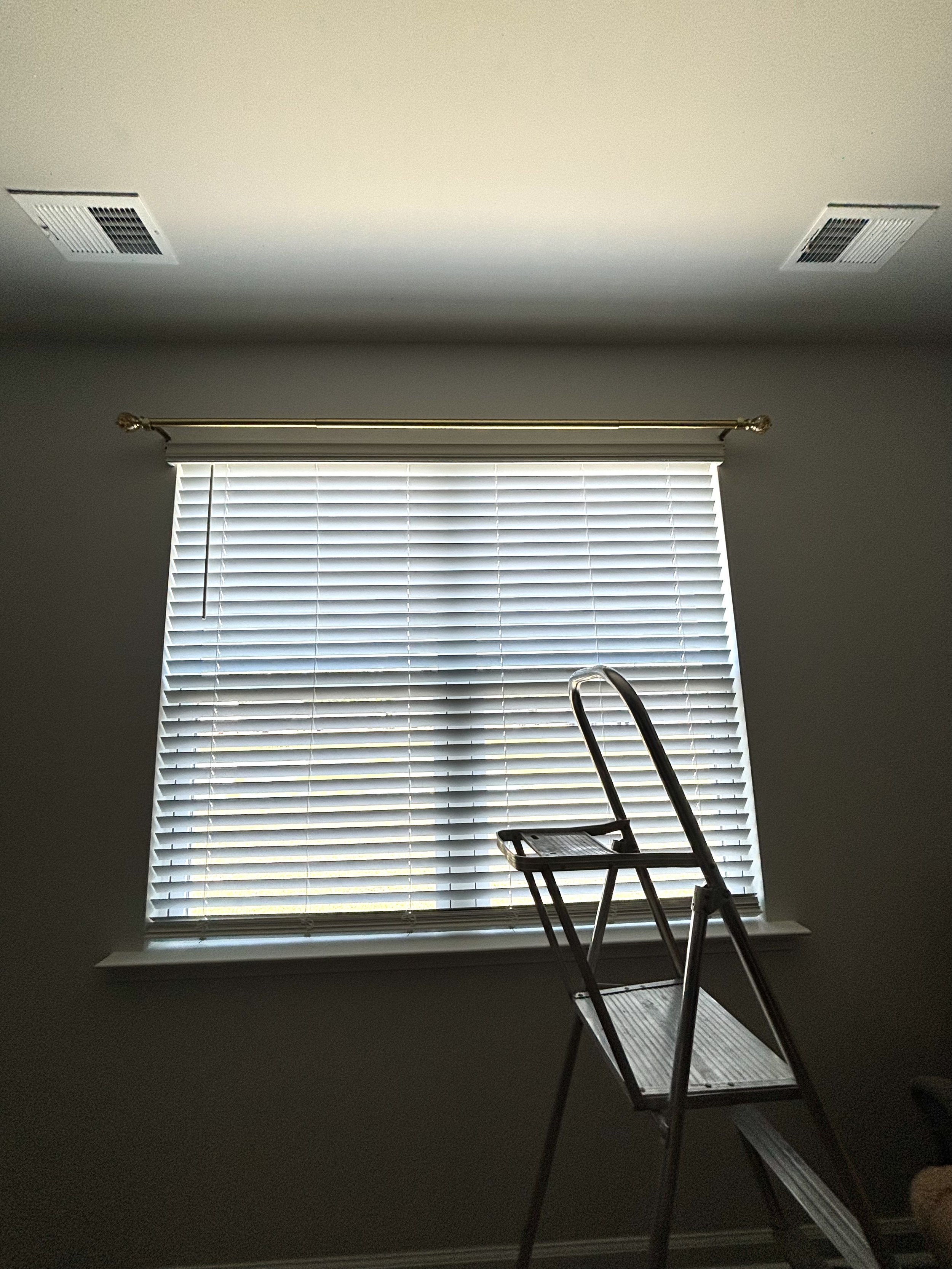 A ladder in front of a window with closed white blinds, located in a room with beige walls and ceiling, with two air vents visible on the ceiling.
