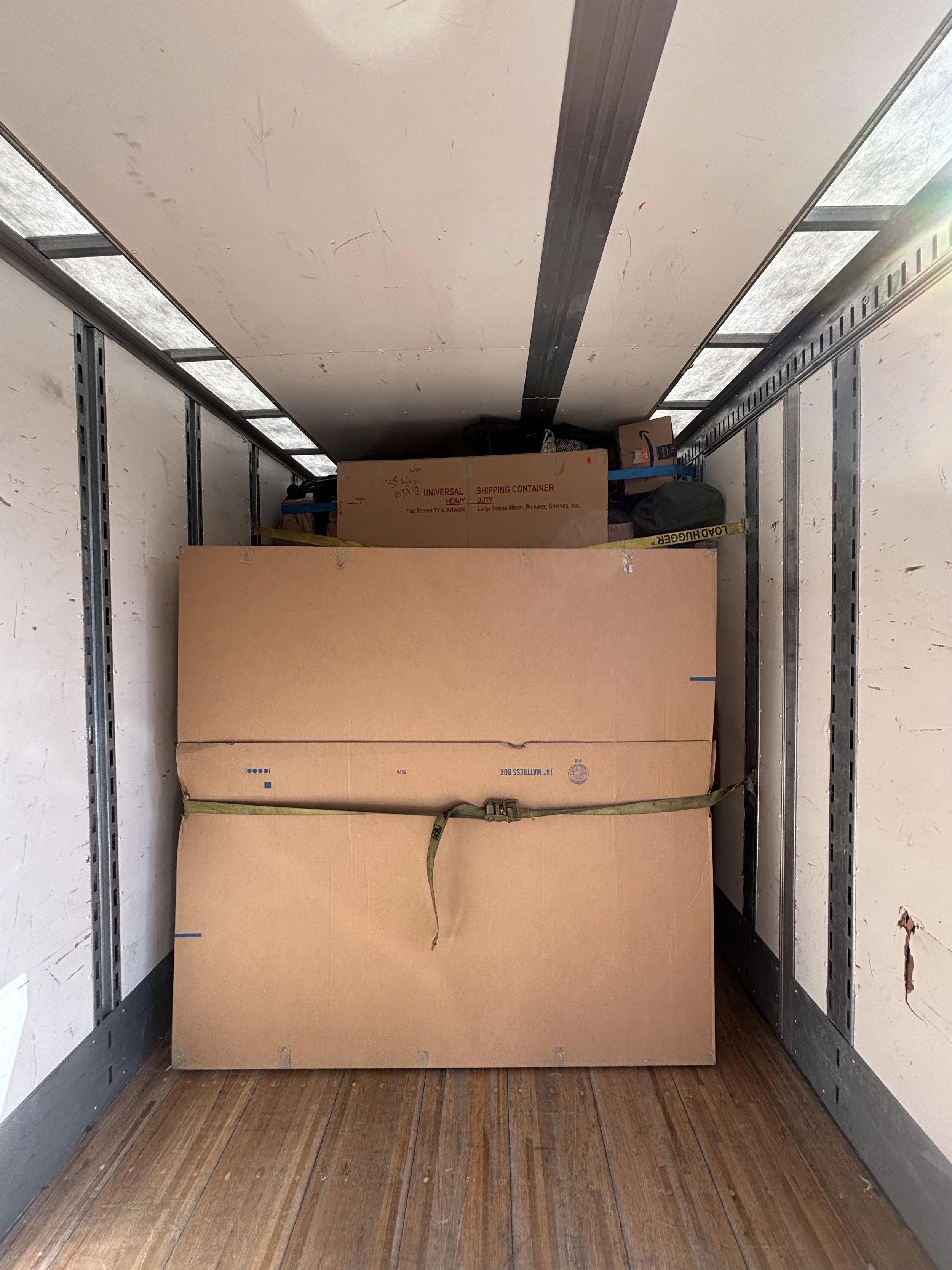 Inside of a moving truck with a large cardboard box secured with green straps on a wooden floor.