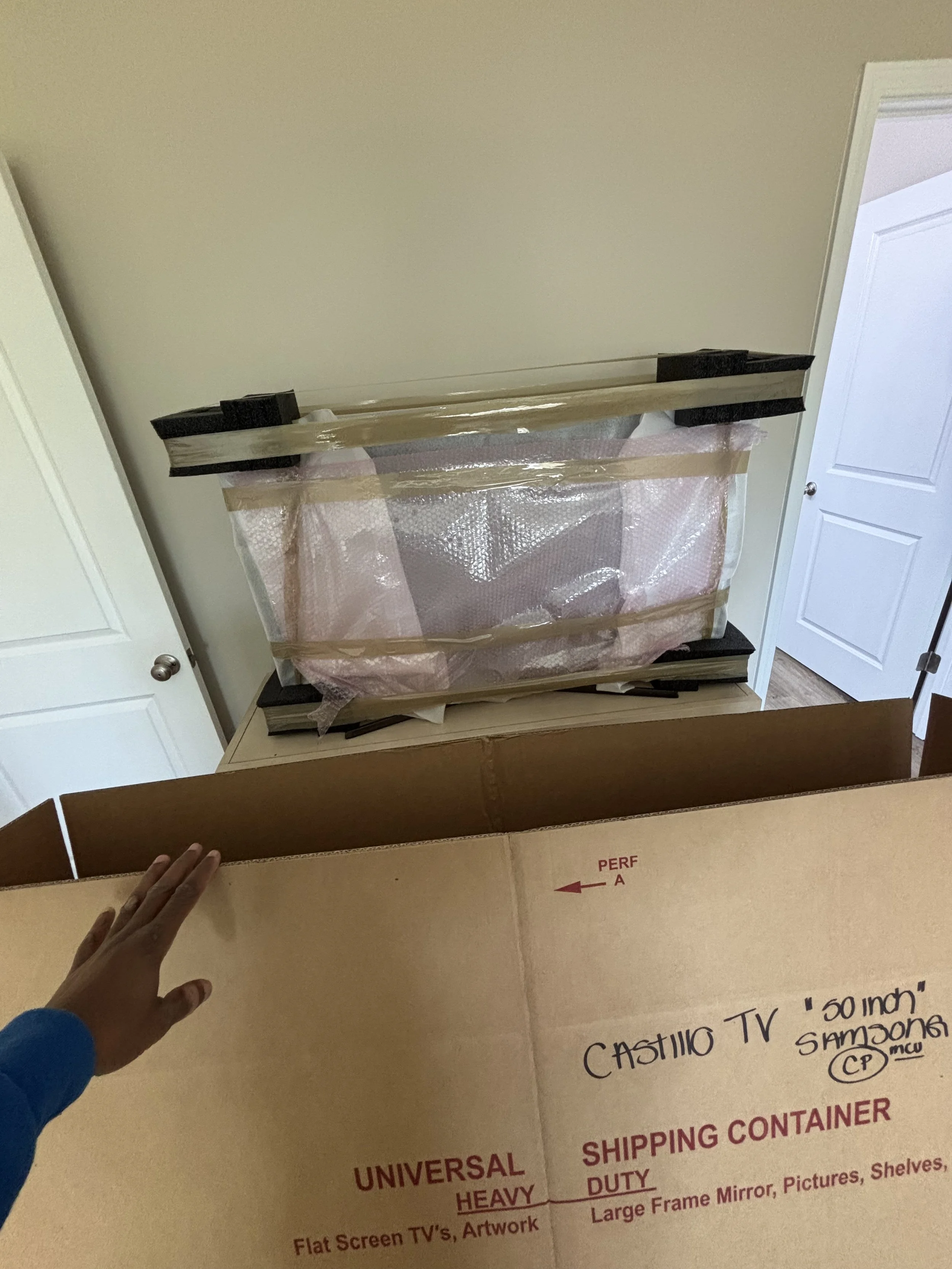 A person wearing a blue long sleeve shirt is holding a large cardboard box labeled 'Castillo TV 50 inch' with additional text about shipping and packaging.