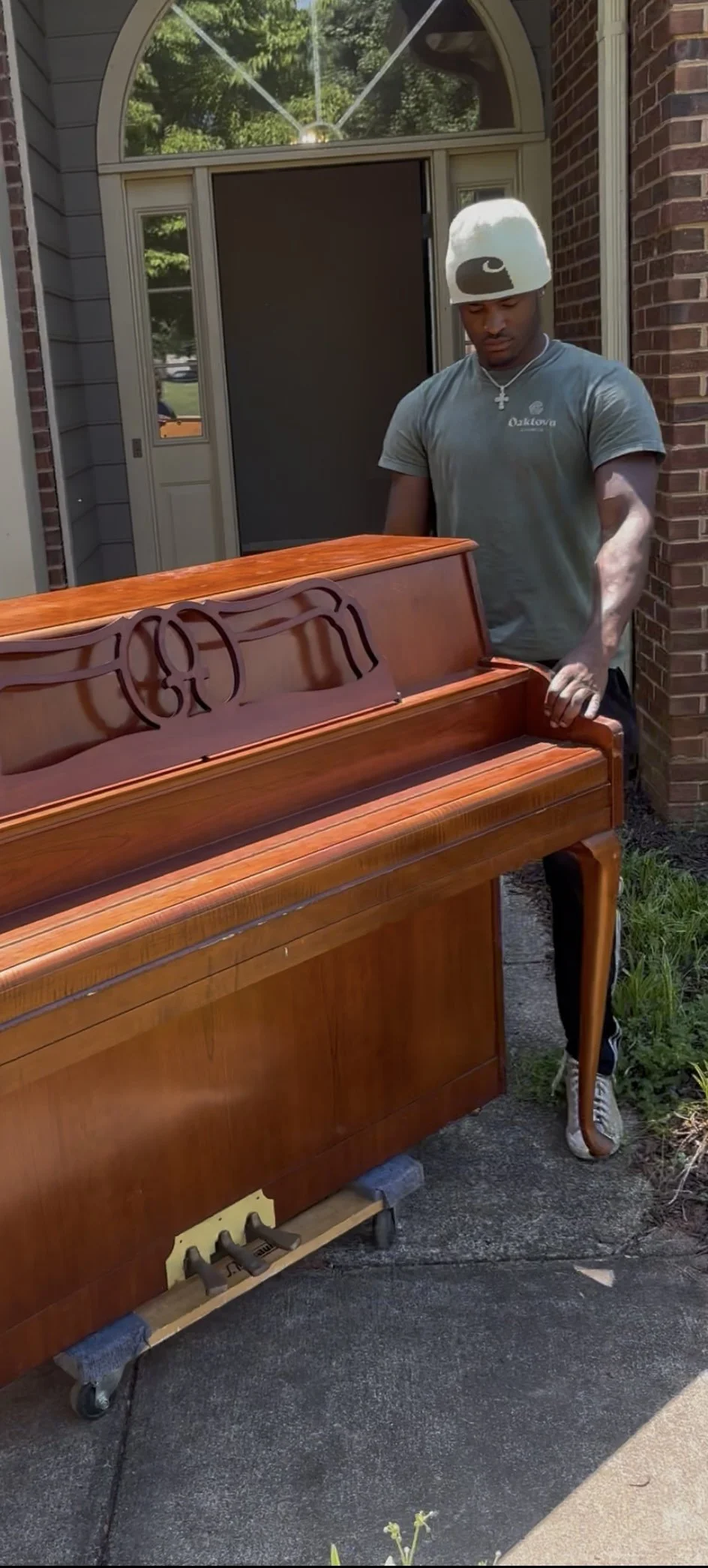 Young man standing outside, looking at a wooden upright piano on wheels, positioned on a sidewalk near a brick house.