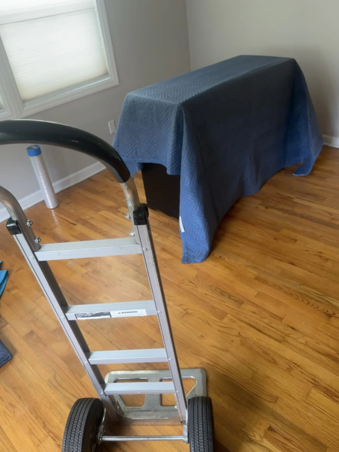 A room with hardwood floors and light-colored walls. A stepladder is in the foreground. In the background, there is a table or piece of furniture covered with a dark blue quilted cover. A window with white blinds is on the left side.