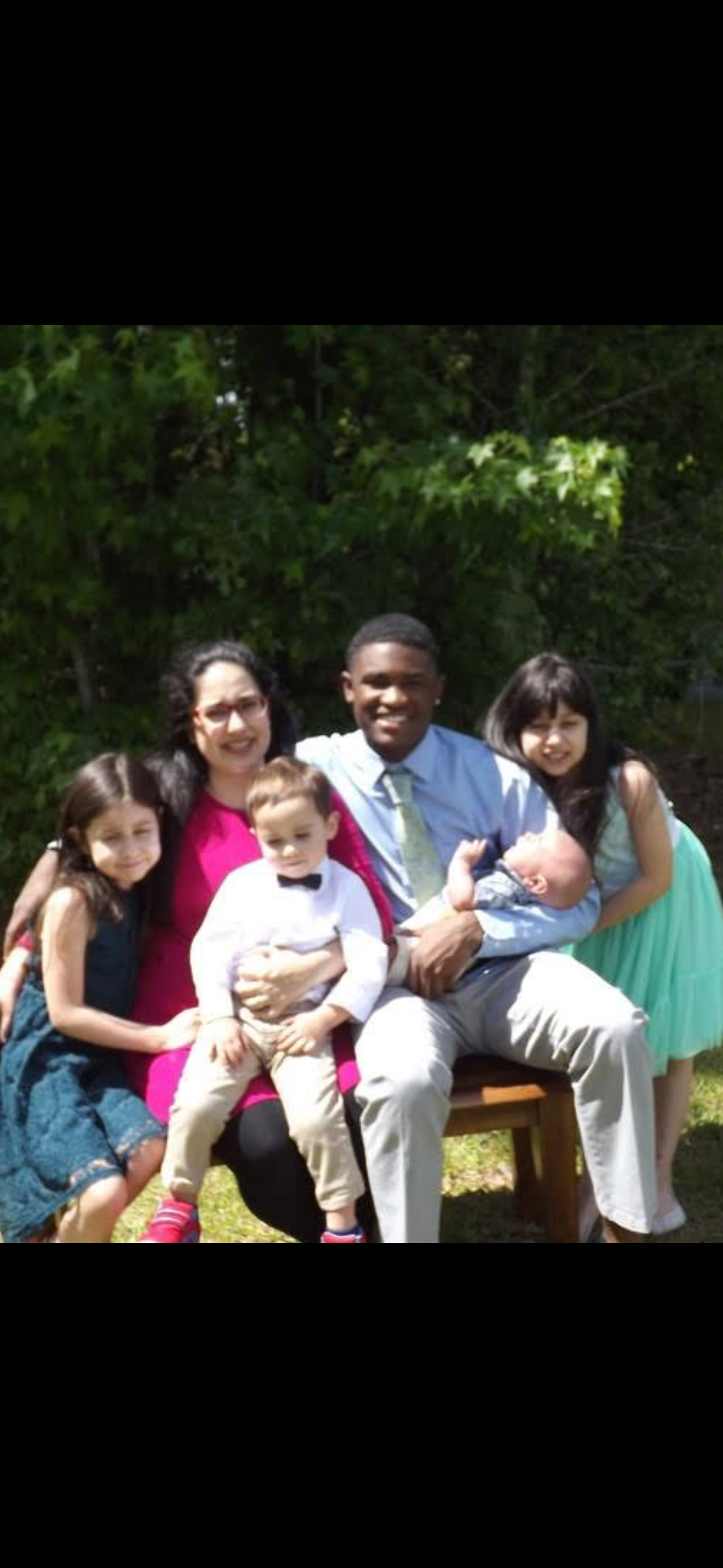 A multicultural group of six children and two adults sitting outdoors on a bench, with trees and greenery in the background, smiling for the photo.