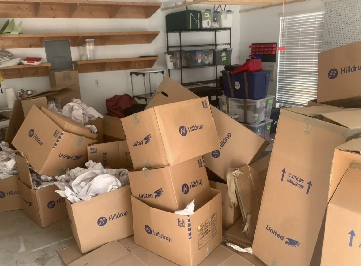 A cluttered room filled with stacked cardboard boxes labeled 'Hilldrup' and 'United' and crumpled packing paper, with shelves and storage containers against the walls.