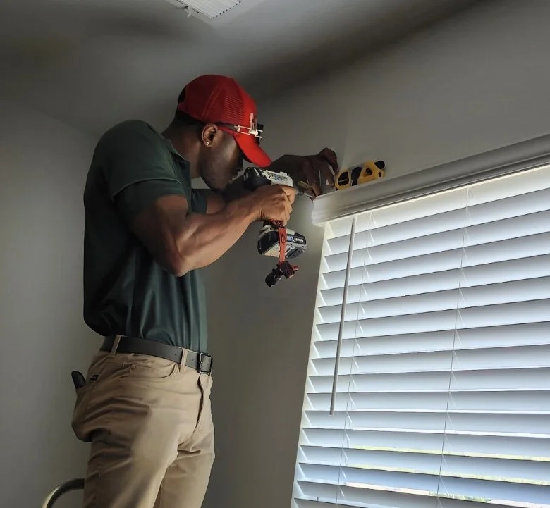 A man in a green polo shirt, beige pants, and a red baseball cap is using a drill to work on a white window blind at the top of a window.