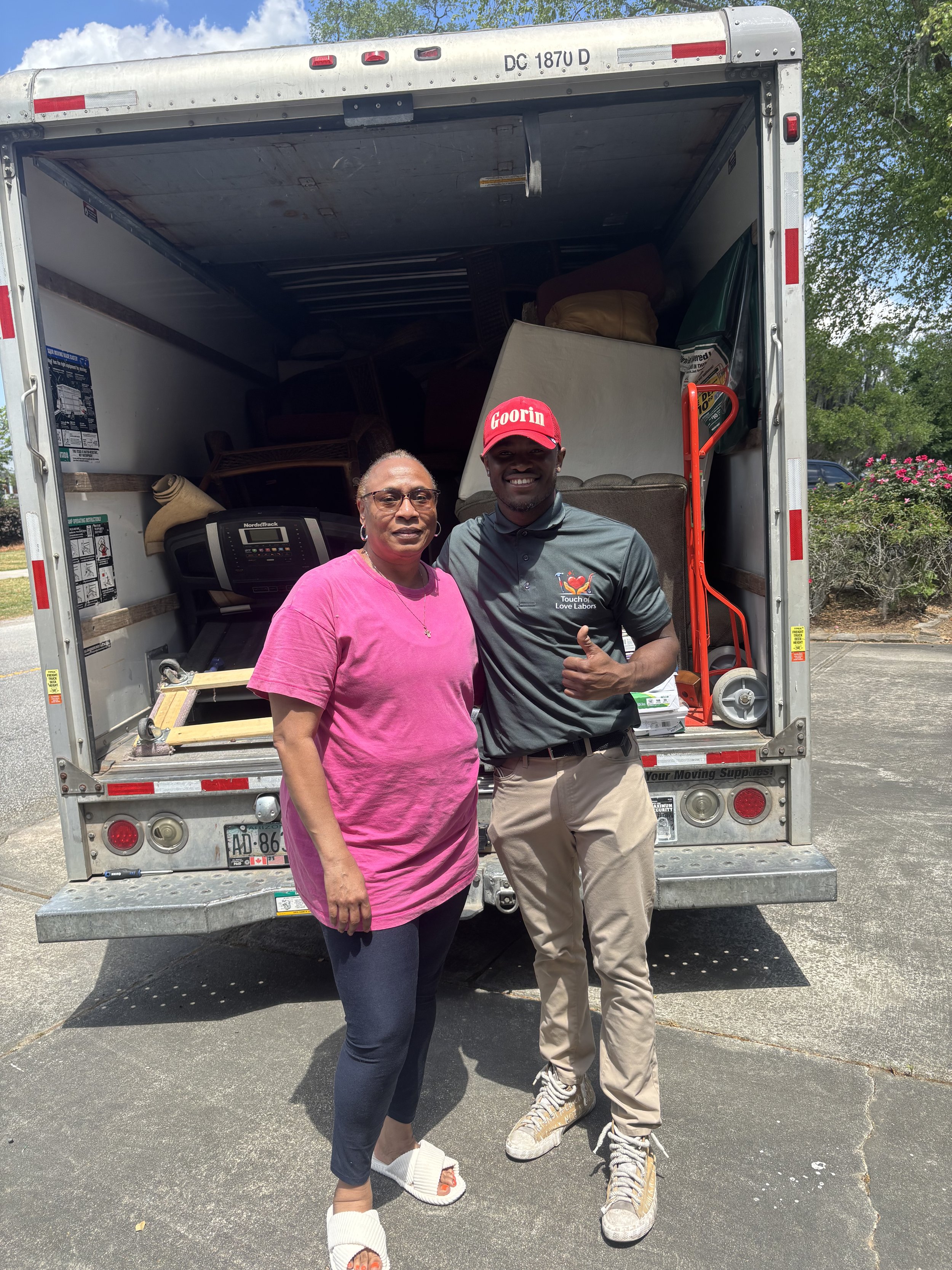 Two women stand in front of an open moving truck. One woman is wearing a pink shirt and black leggings, and the other is wearing a gray polo shirt, khaki pants, and large sneakers. The woman in the polo shirt is giving a thumbs-up and smiling. The truck appears to be loaded with furniture and boxes.