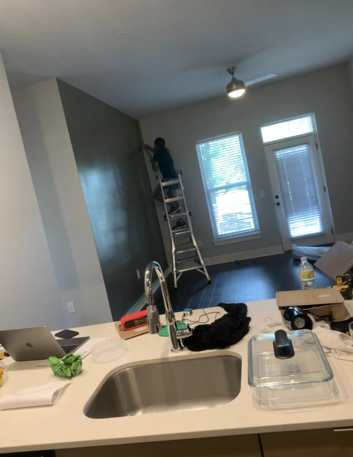 A person standing on a ladder painting a wall in a living room with large windows and a sliding door. The foreground shows a kitchen island with various items, including a laptop, water bottle, and tools.