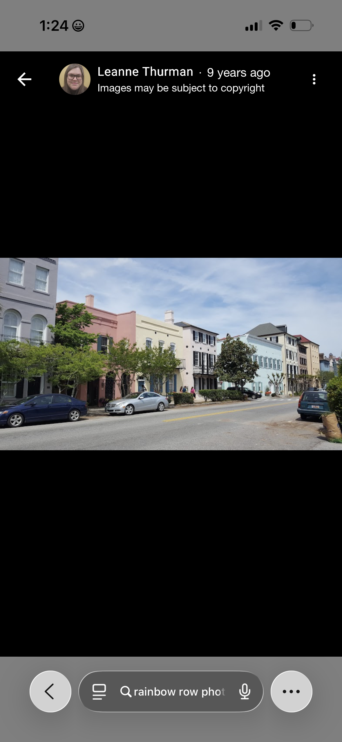 A street with colorful buildings, parked cars, trees, and people walking on sidewalks under a partly cloudy sky.