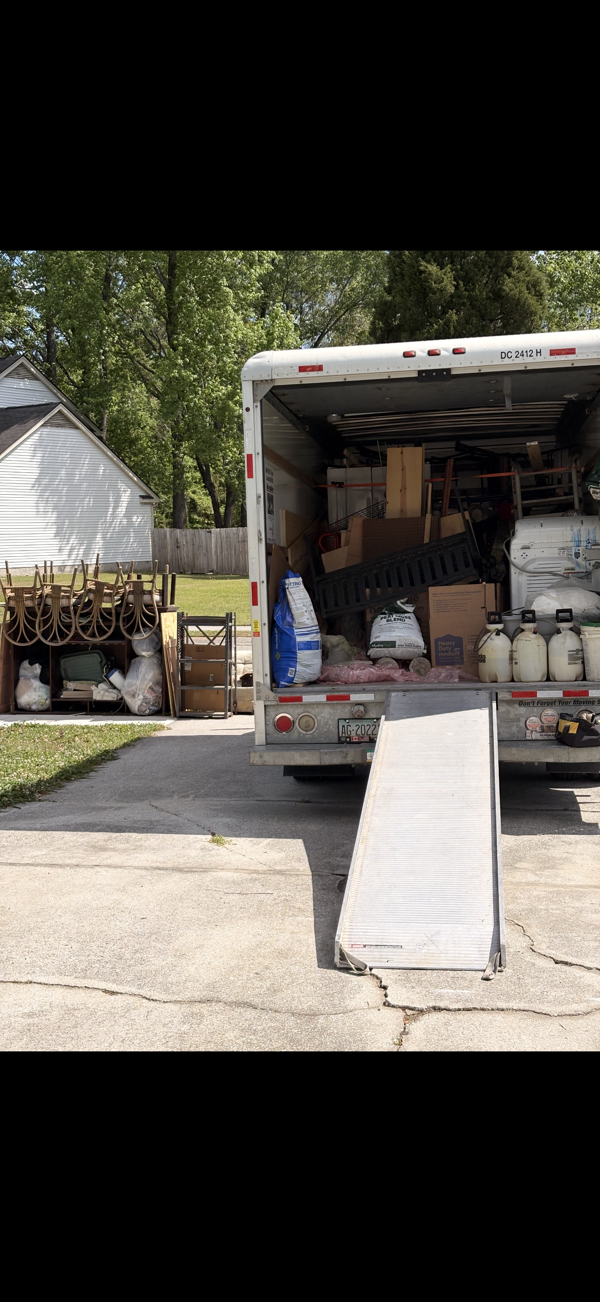 Open moving truck filled with various household items, with a ramp extended to the driveway. To the left, a pile of chairs, boxes, and bags are stacked near a wooden staircase. In the background, a house with a white exterior and a wooden fence surround a yard with trees.