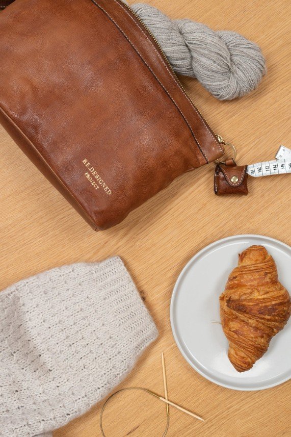 Vaalea puiset pöytä, jossa kello ja pieni ruskea nahkalaukku, harmaa villapaita, harmaa lanka ja mittanauha, valkoinen lautanen ja croissant.