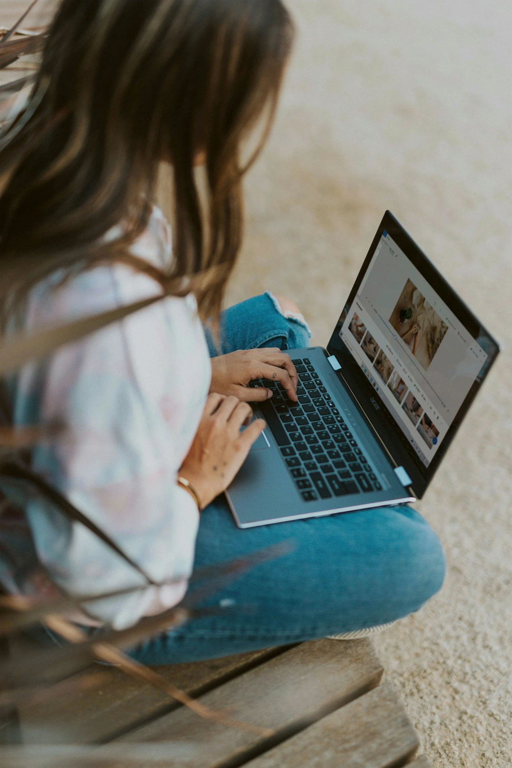 A brunette woman wearing jeans and a shirt sits outside with a laptop on her knee.