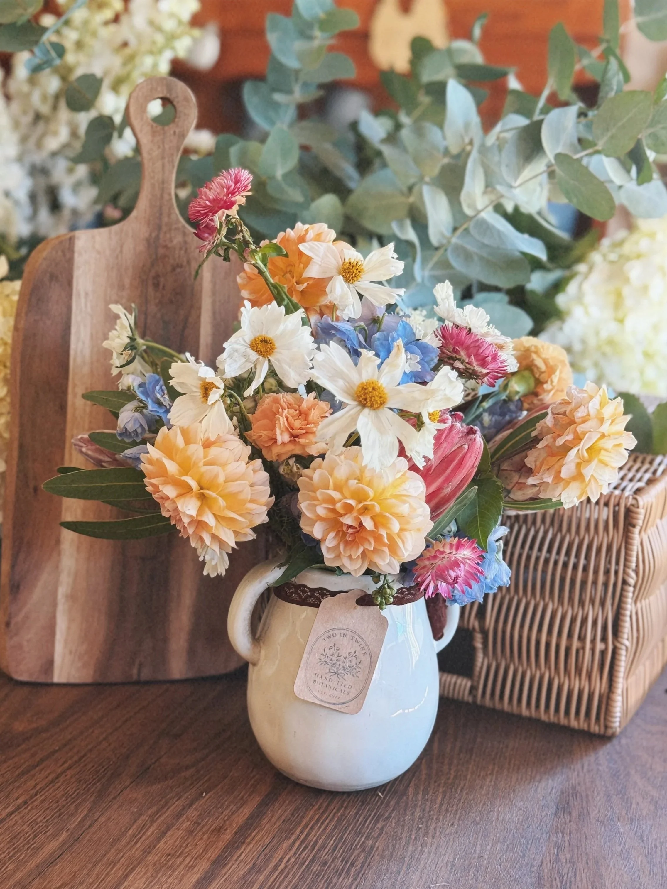 A bouquet of mixed flowers including white daisies, pink carnations, peach roses, and blue delphiniums arranged in a white ceramic pitcher with a vintage-style tag hanging from the handle. The pitcher is placed on a wooden table with a wooden cutting board and a wicker chair in the background.