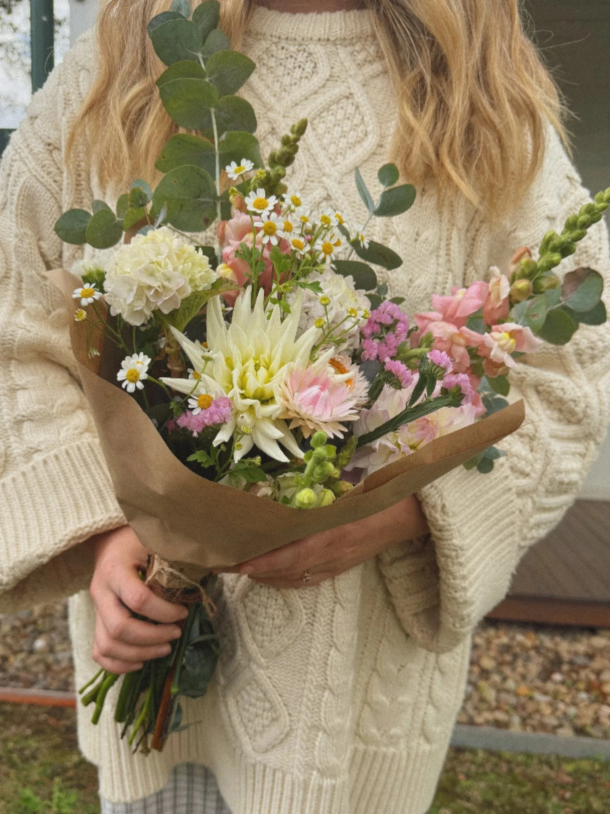 Person in cream-colored cable-knit sweater holding a bouquet of mixed flowers with greenery outdoors.