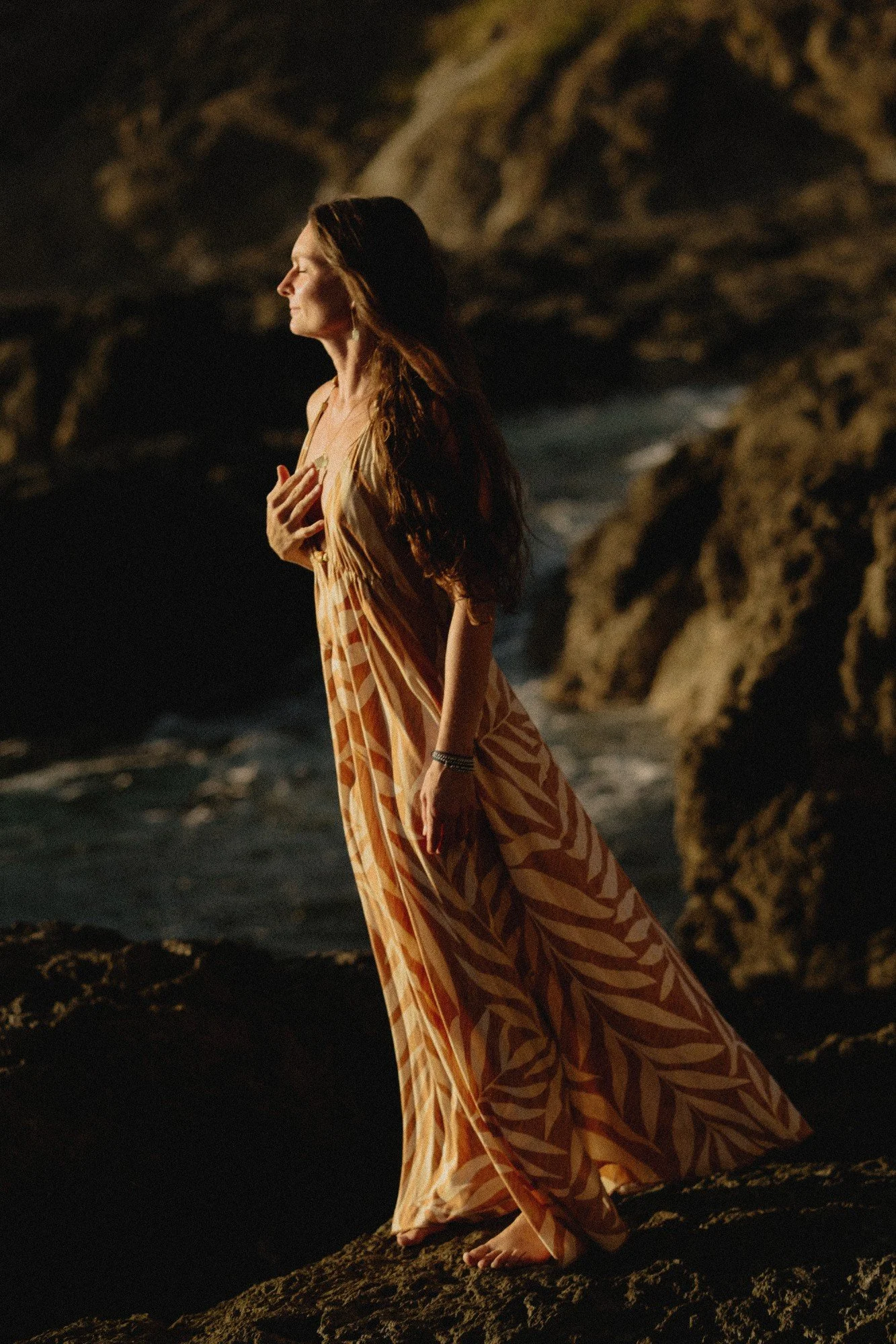 A woman with long hair wearing a patterned orange and yellow dress stands on rocks near the ocean at sunset, with her eyes closed and hand on her chest.