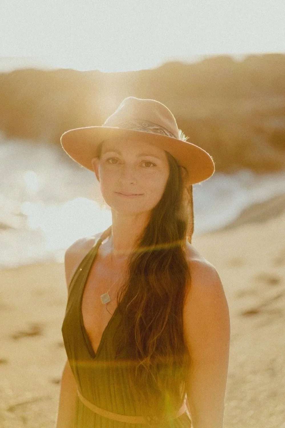 Woman smiling on a beach at sunset, wearing a wide-brimmed hat and a sleeveless top.