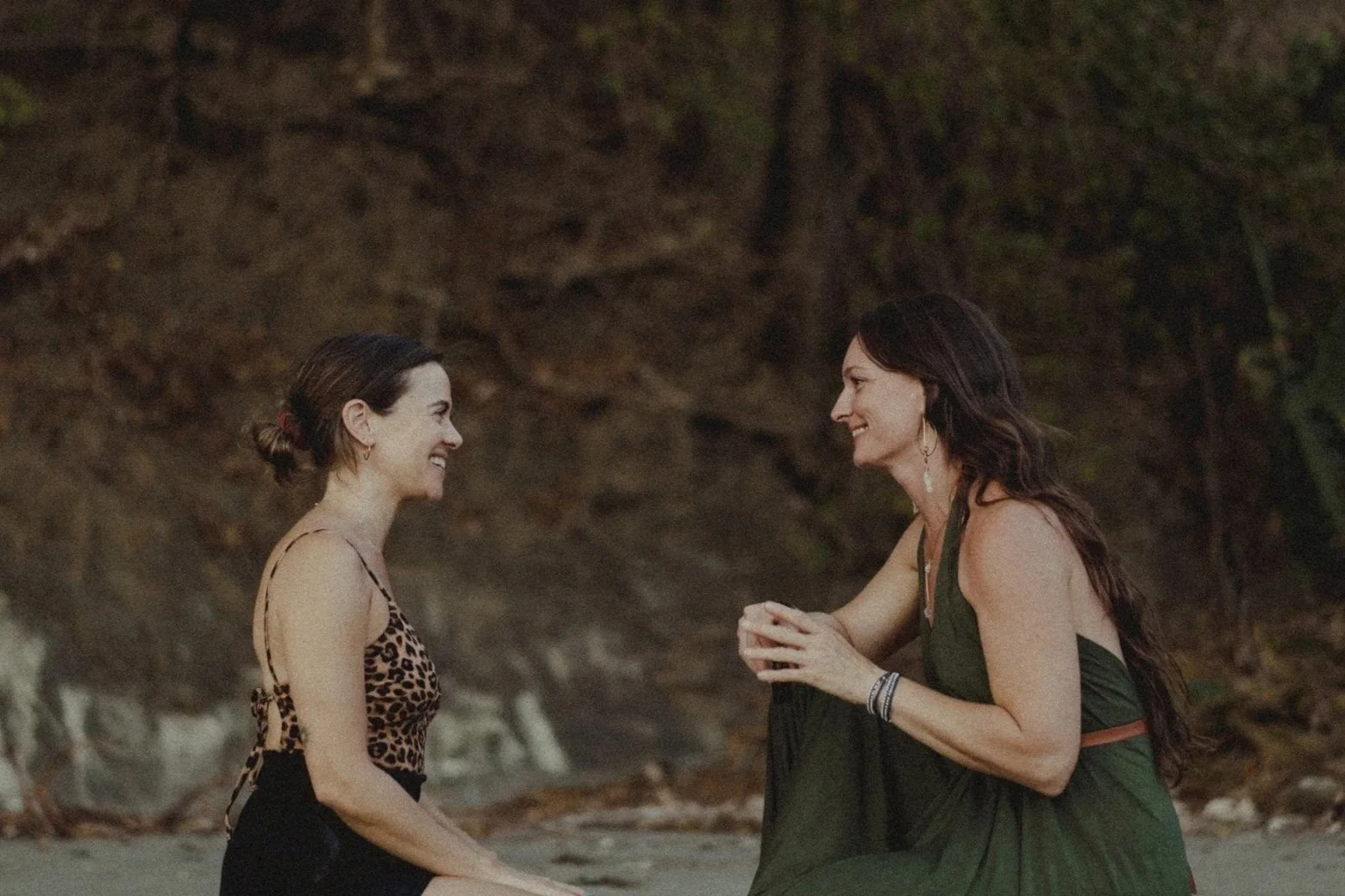 Two women smiling and sitting facing each other on a beach with a rocky cliff behind them. One woman is in a leopard print top, and the other is in a dark green dress with earrings.