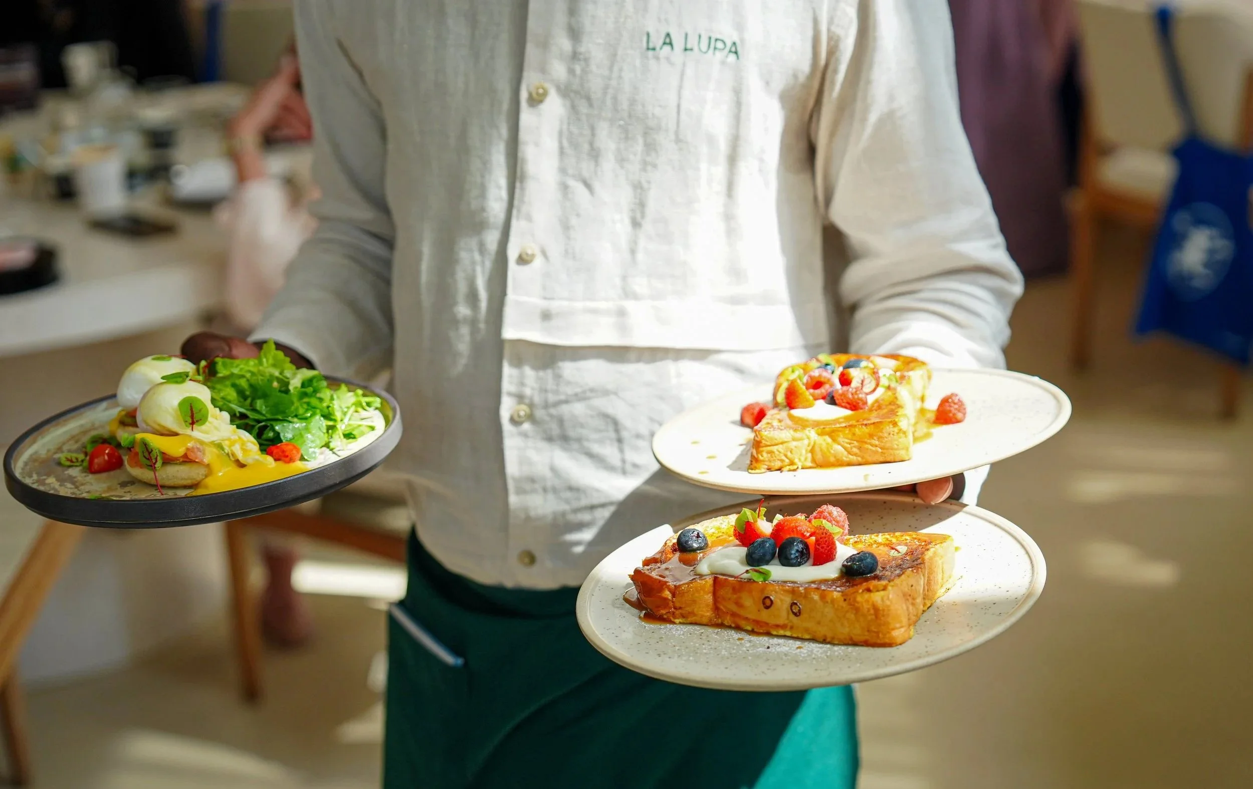 Person holding three plates with salads and desserts, in an indoor setting.