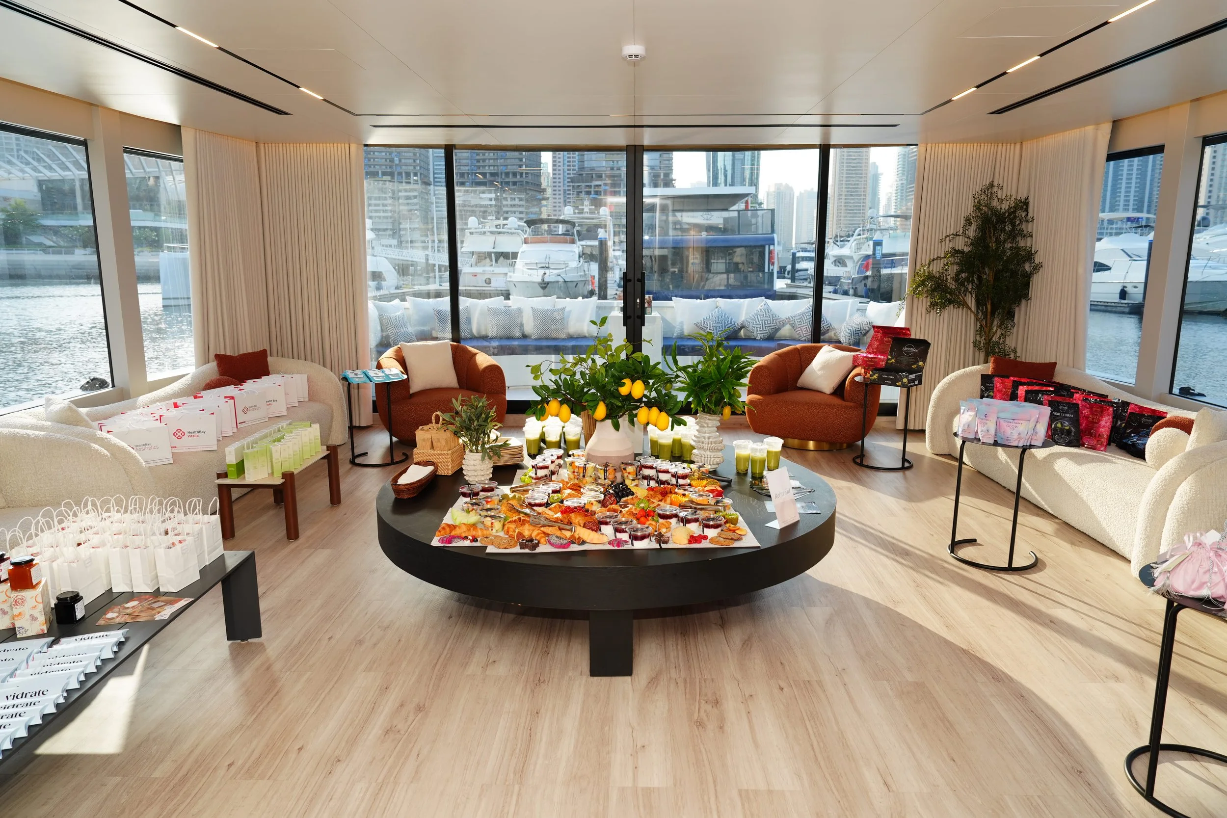 Luxury yacht lounge with a brunch spread, citrus centerpiece, gift bags, and marina views of boats and skyscrapers
