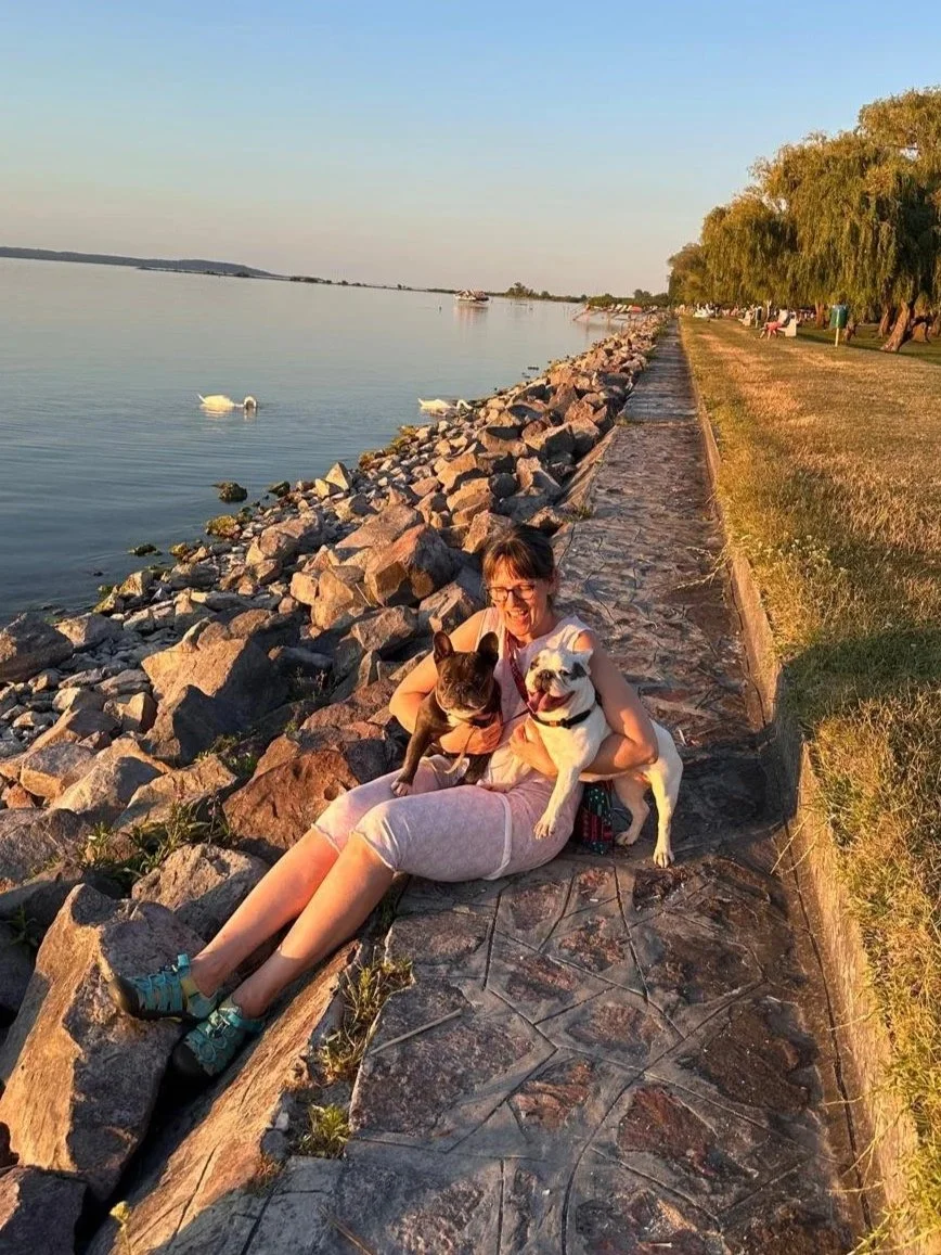 A woman sitting on a rocky pathway by a lake, holding two Frenchies, with ducks swimming in the water nearby. The setting appears to be during sunset with warm lighting.