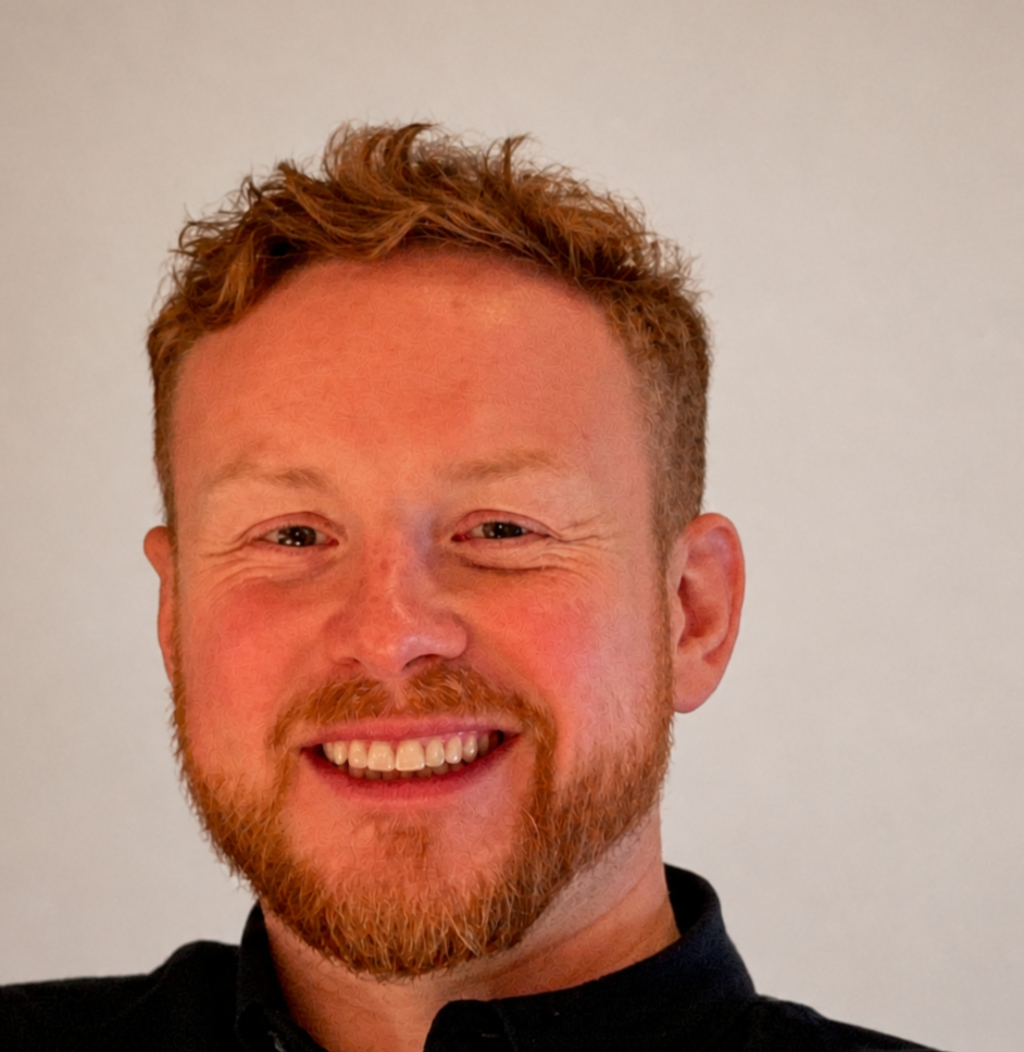 A smiling man with short, curly red hair and a beard, wearing a black shirt, standing against a plain light-colored background.