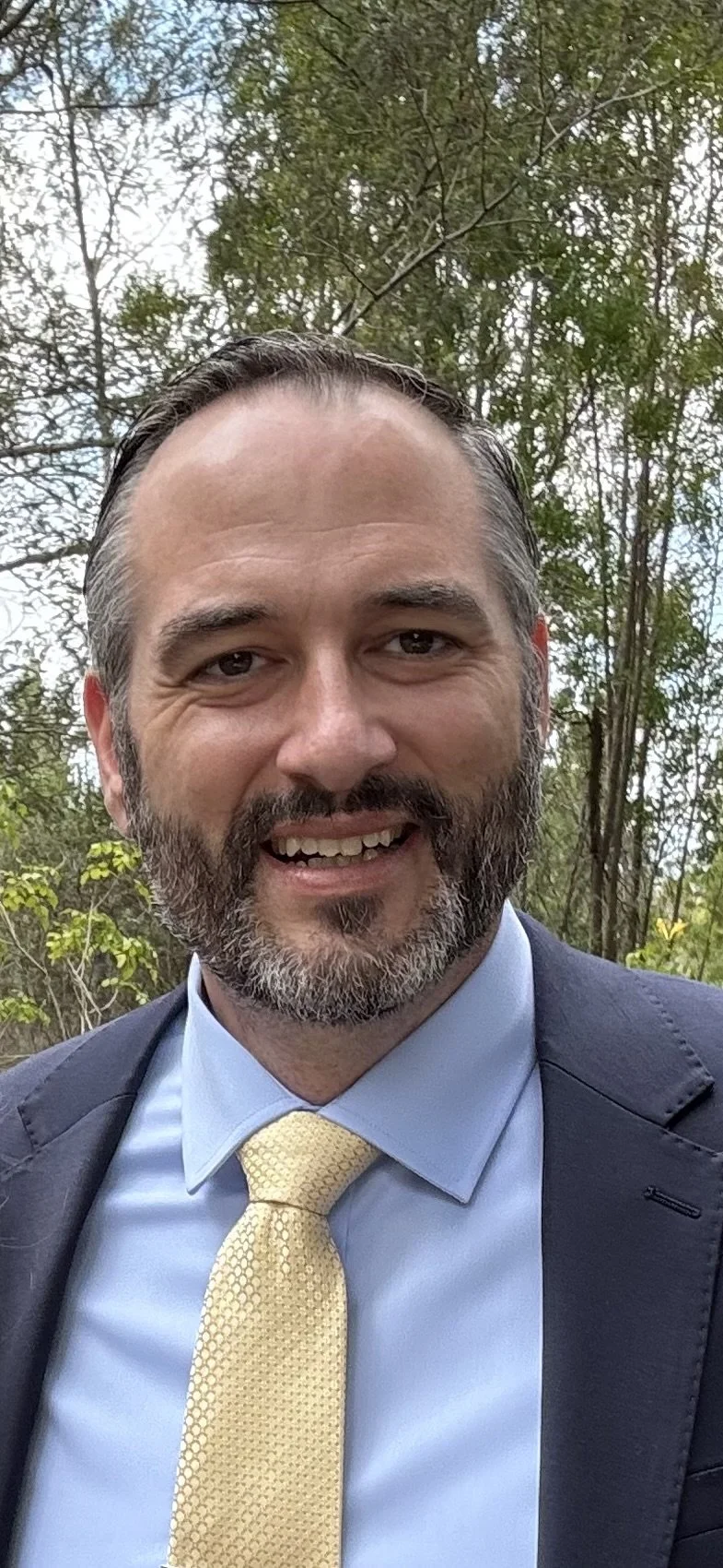 Man smiling outdoors, dressed in a blue shirt, yellow tie, and dark blazer, with trees in the background.