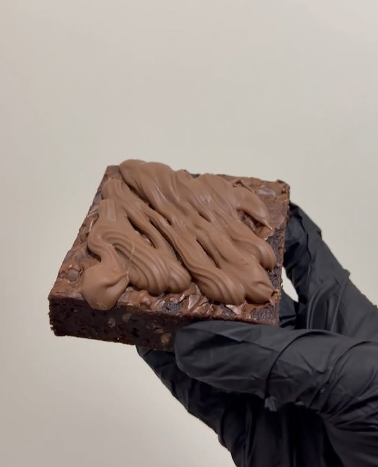 A hand wearing a black glove holding a chocolate brownie with chocolate frosting on top against a plain, light background.